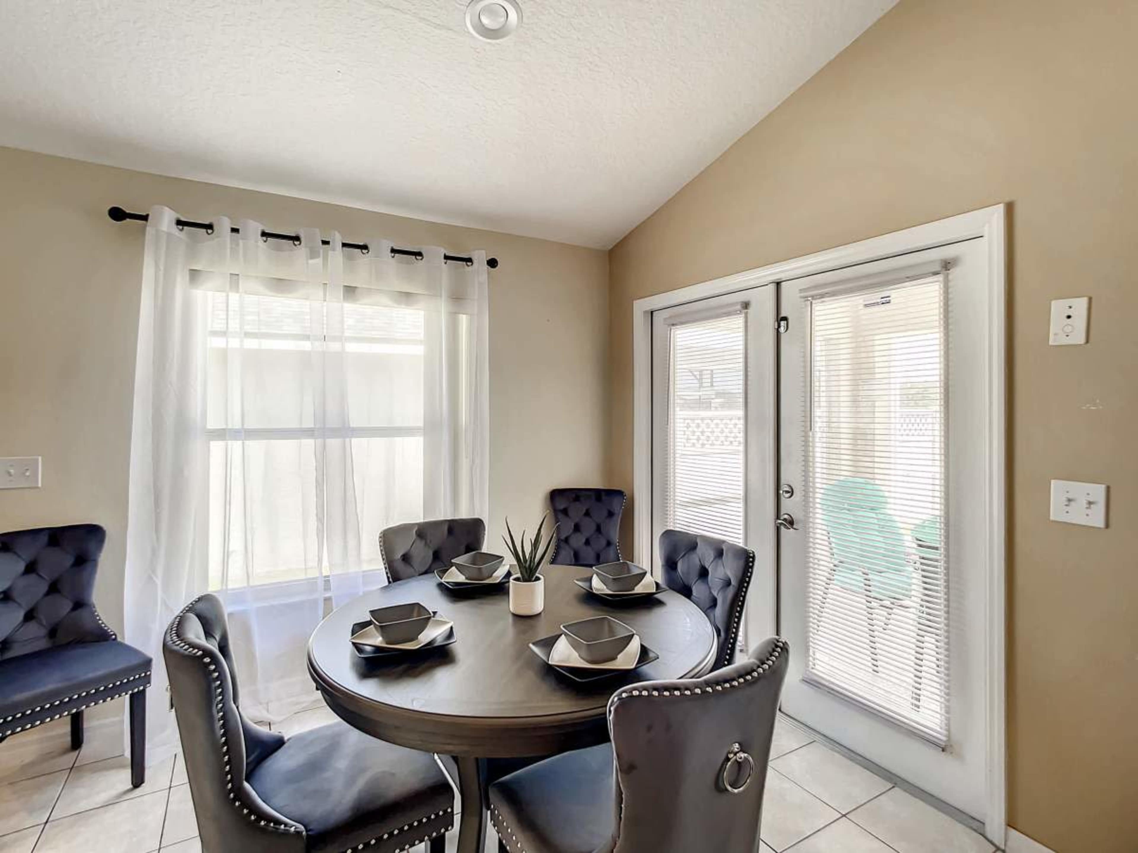 A round dining table is set with four place settings in a small, bright room with a pair of glass doors leading outside.