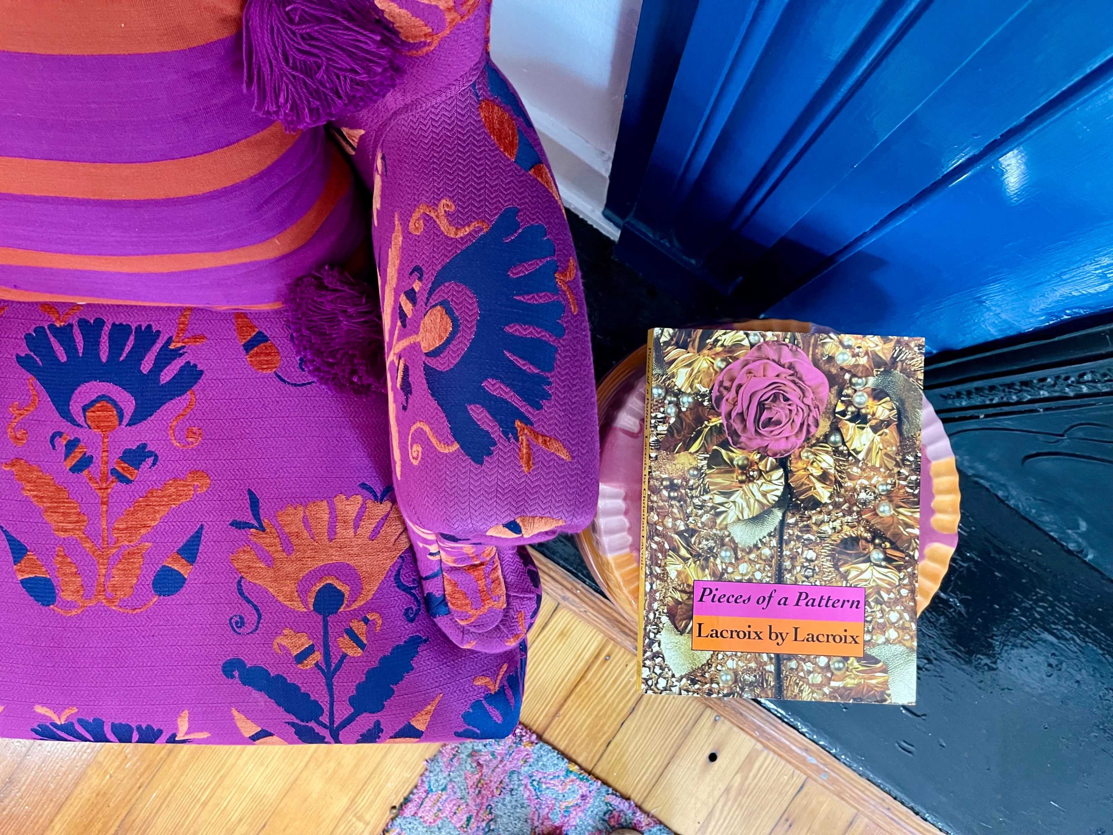 A vibrant patterned chair is paired with a book titled "Pieces of a Pattern: Lacroix by Lacroix," resting on a wooden surface beside a blue wall.