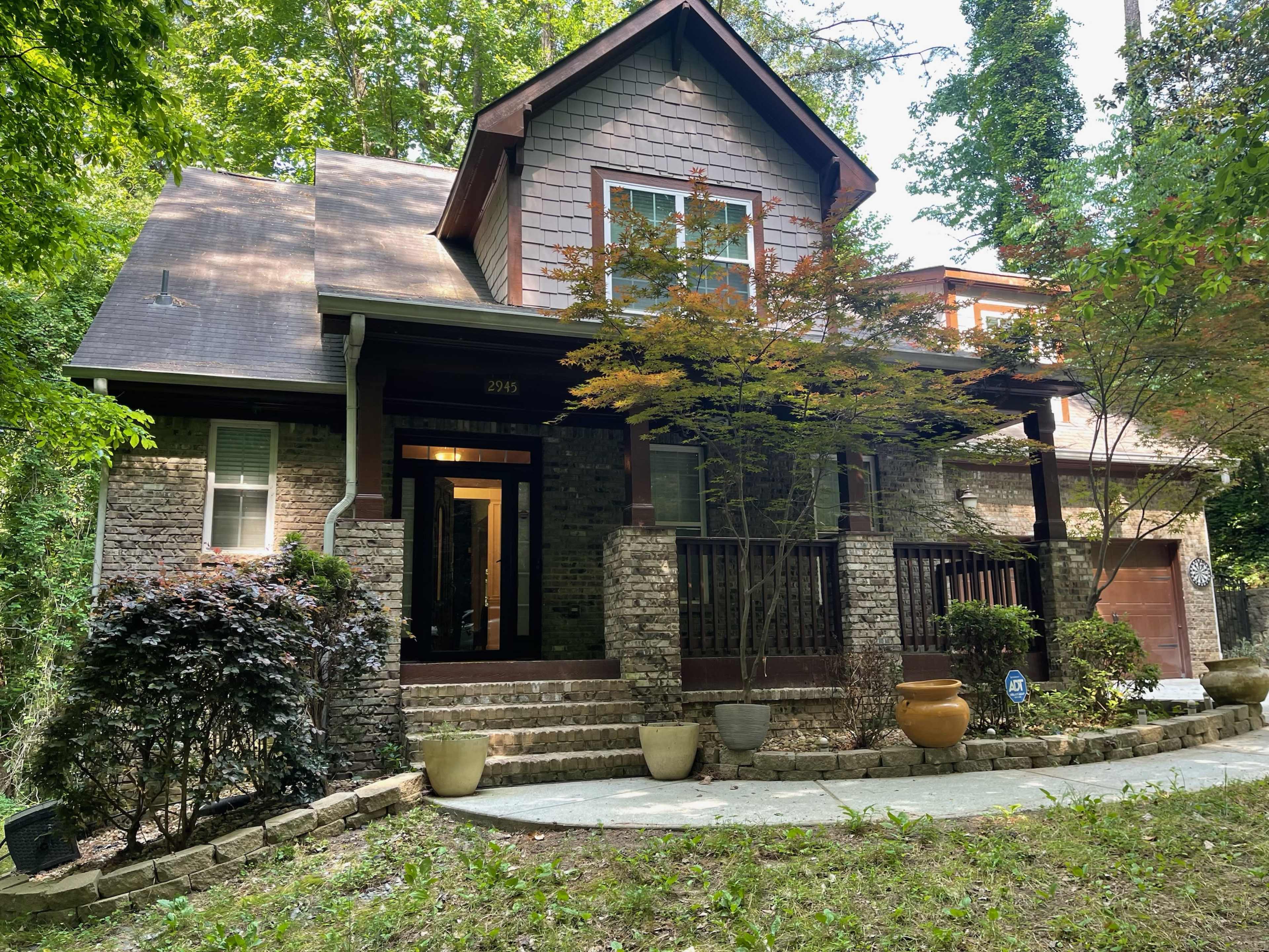 A two-story house with a stone facade and a covered porch is surrounded by greenery and has multiple planters at the entrance.