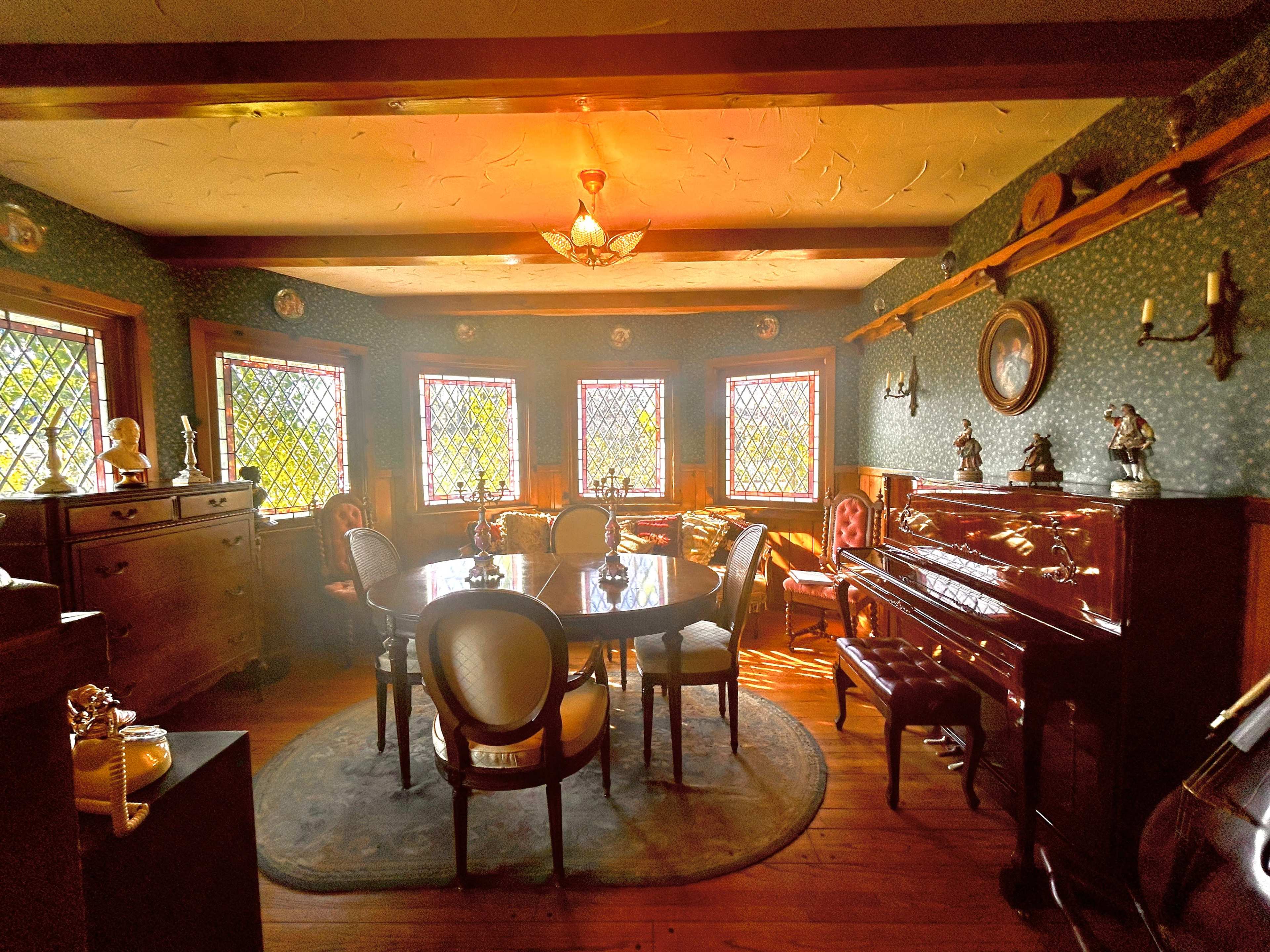 A dining room with a round table surrounded by chairs, illuminated by light filtering through stained glass windows, features a piano and various decorative items.
