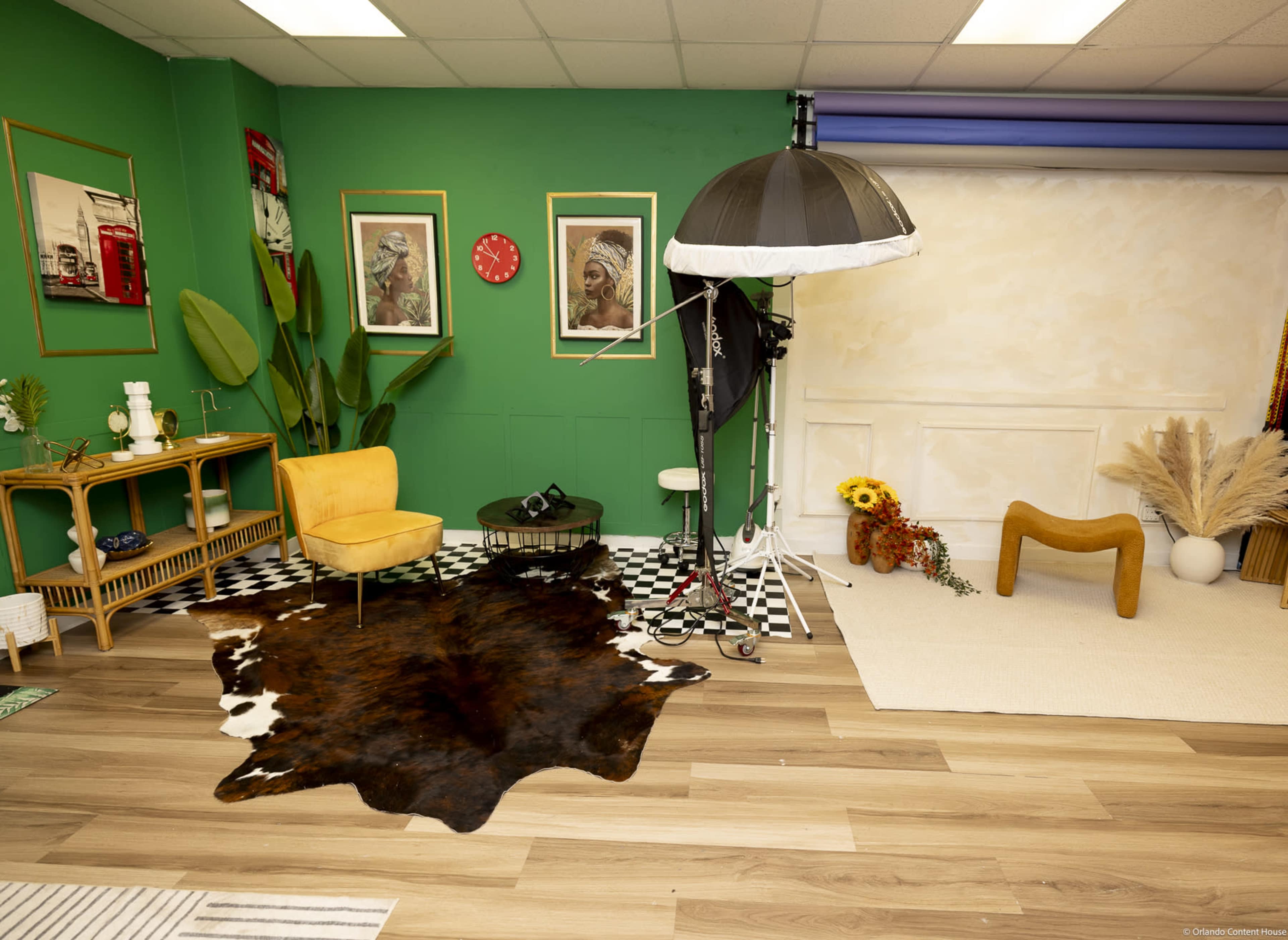 The image shows a photography studio with green walls, a yellow chair, a black and white checkered floor, and various props including a light umbrella and decorative items.