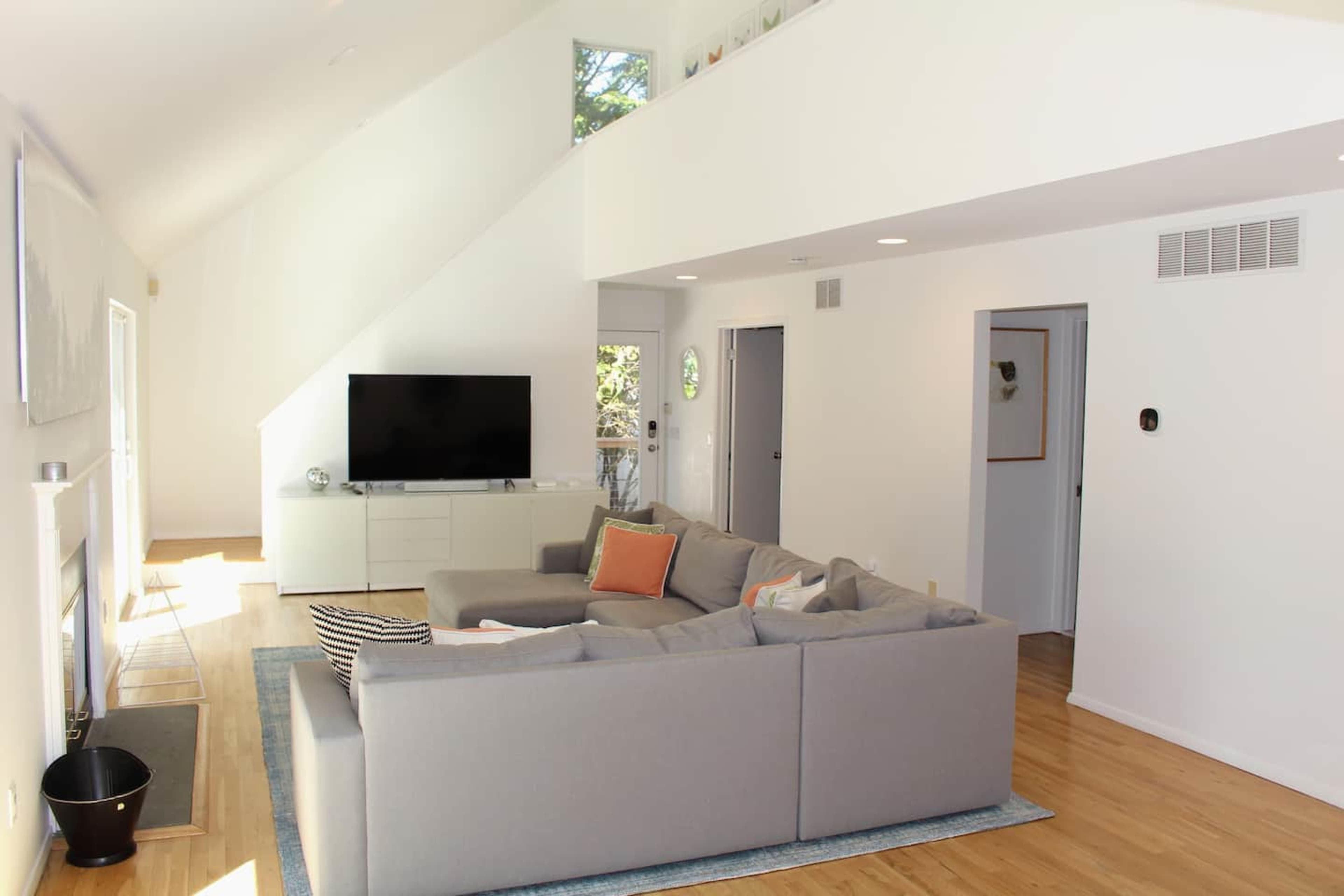 A modern living room with a gray sectional sofa, a television mounted on the wall, and large windows allowing natural light.