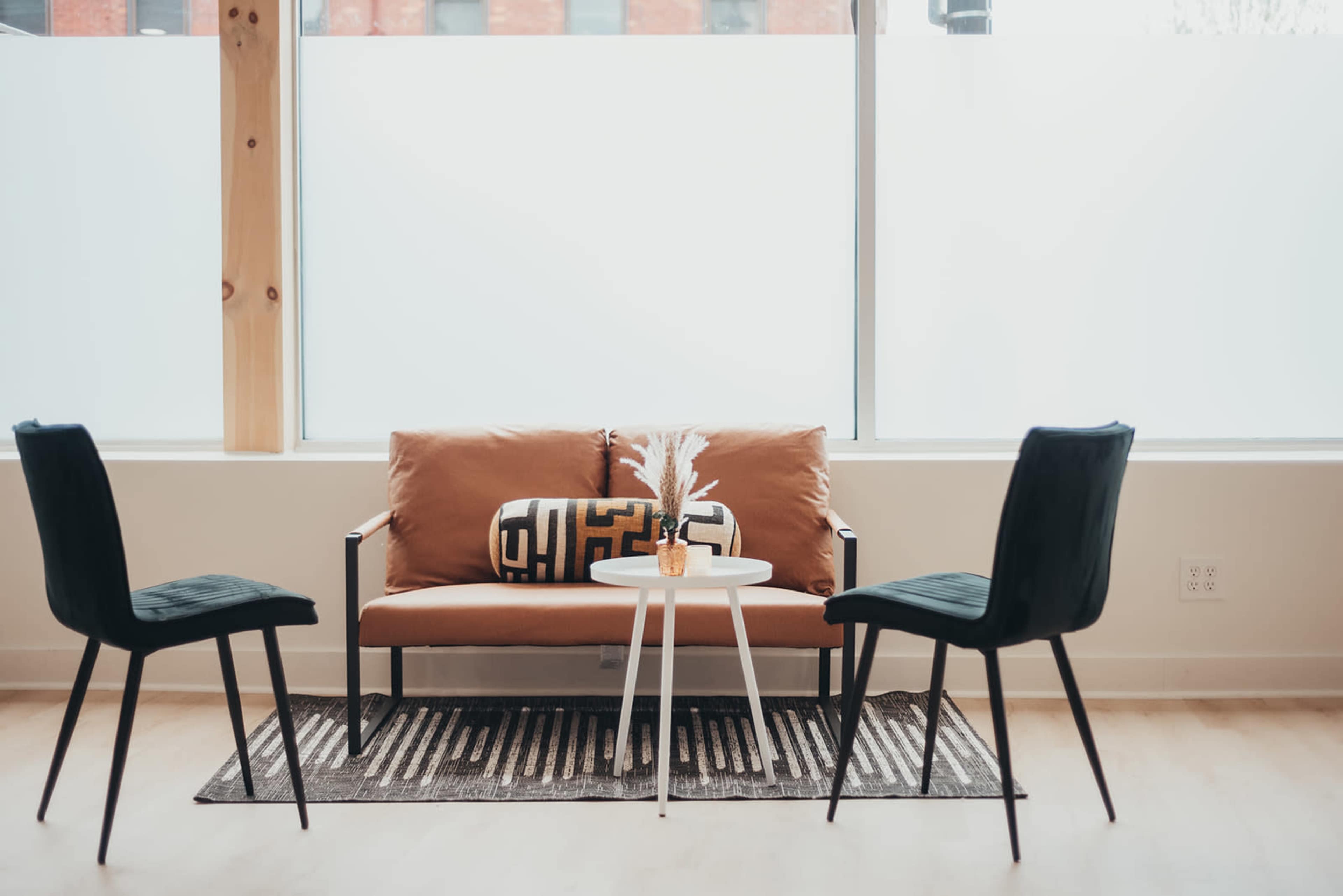 A modern seating area features a brown sofa with a patterned pillow, a round white table, and two black chairs, all set against a large window.