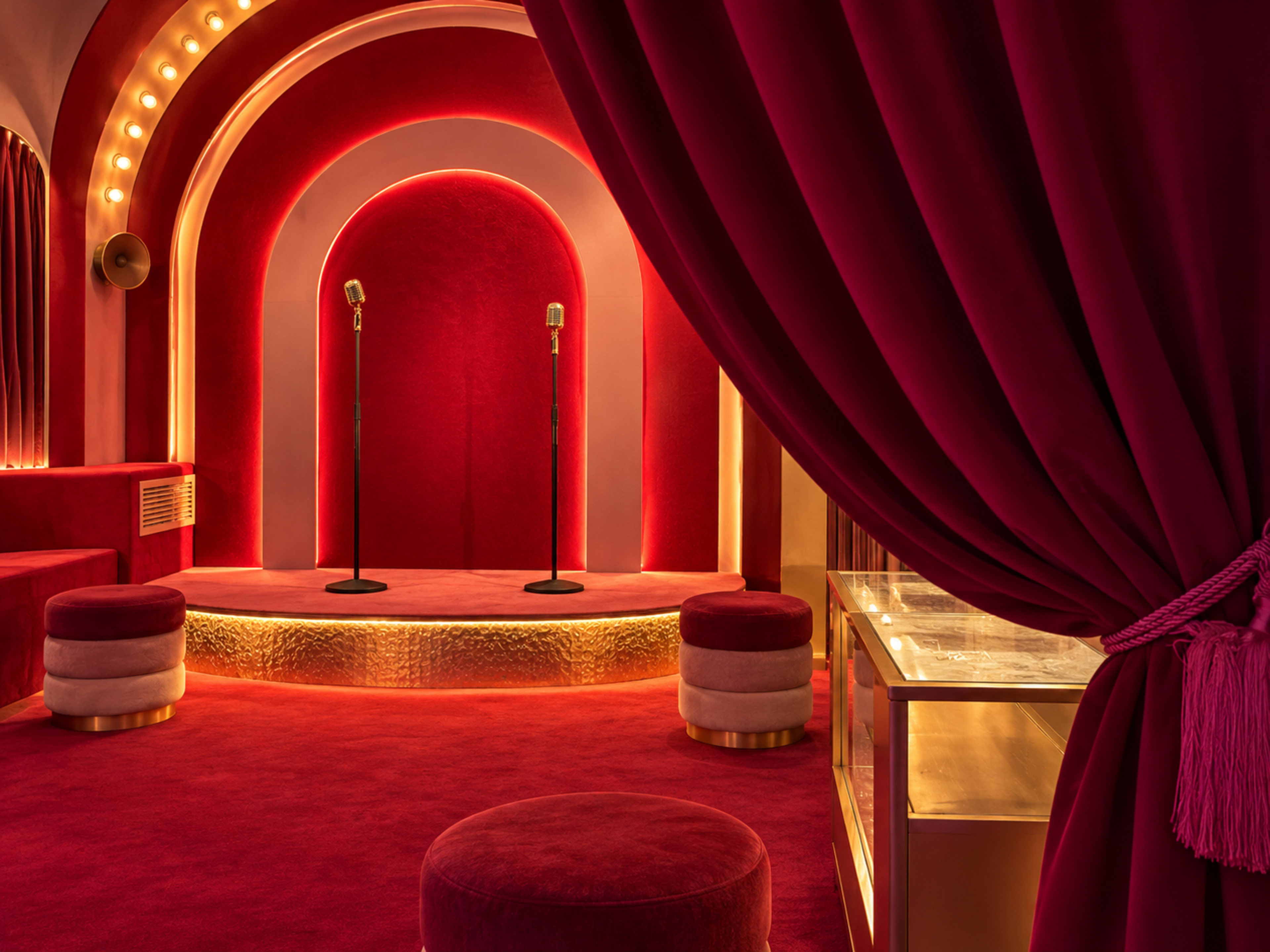 The image shows a brightly lit interior featuring a stage with two microphones, surrounded by deep red velvet drapery and plush seating.