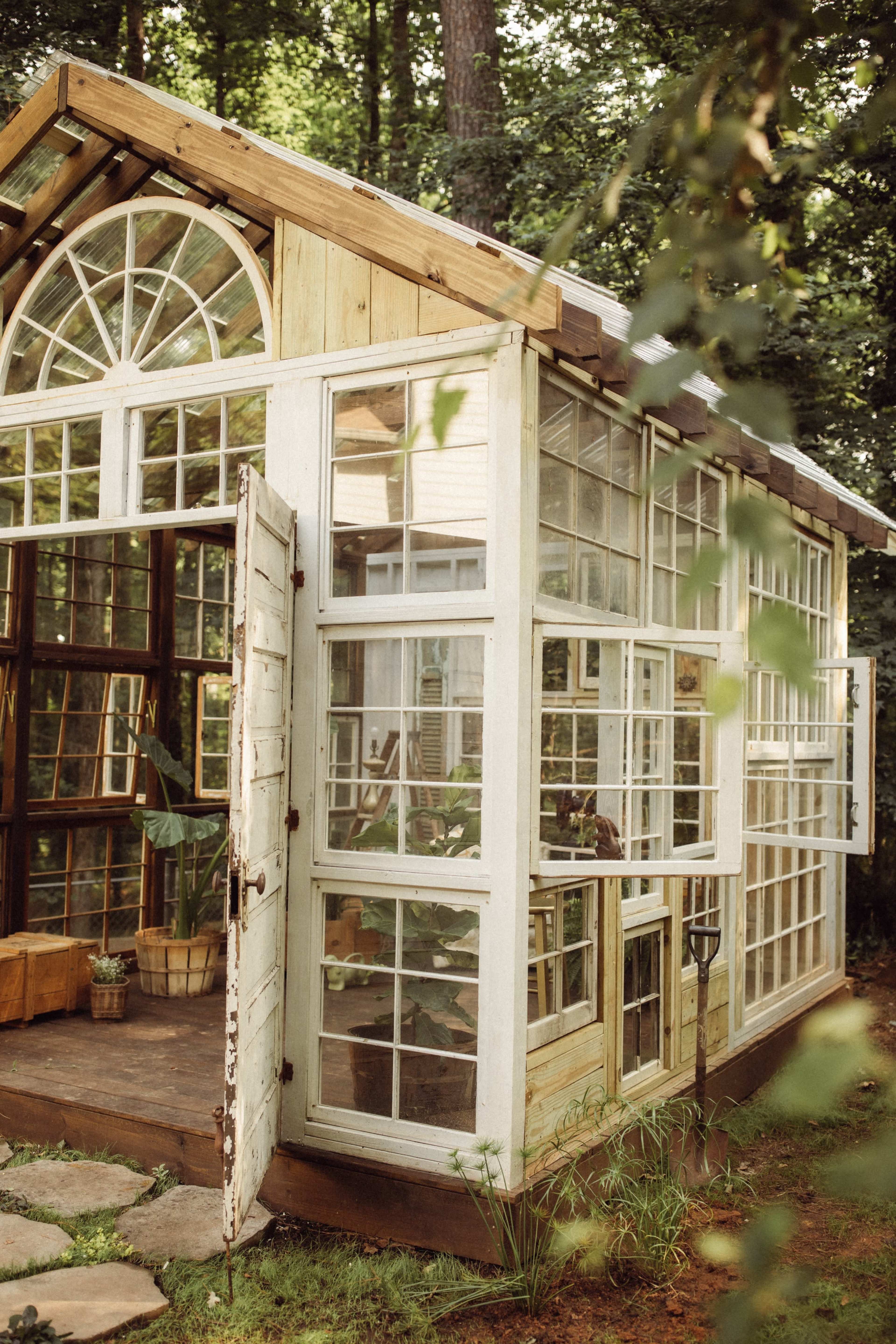 The image shows a wooden and glass greenhouse with multiple panes and an open door, surrounded by greenery.