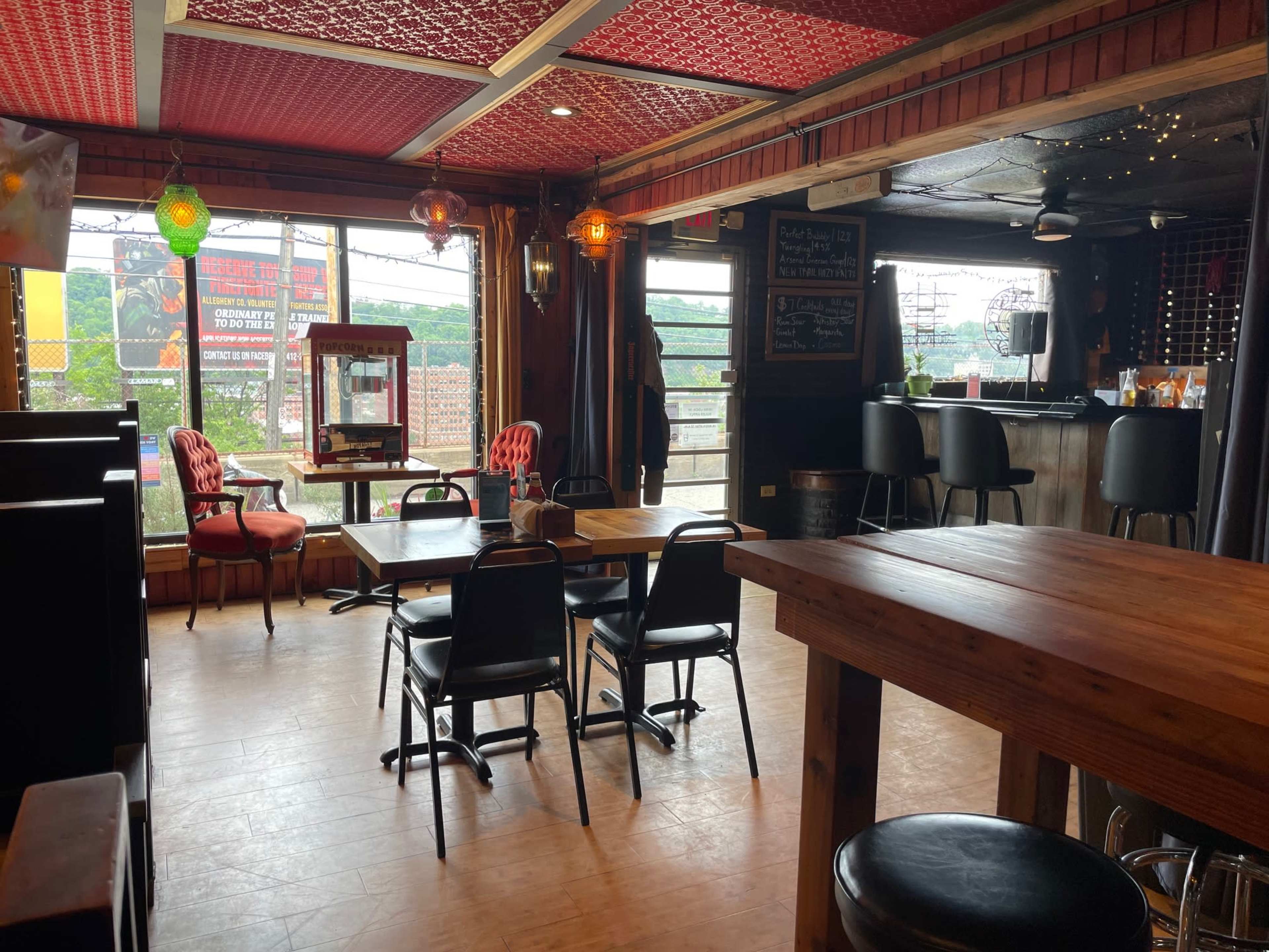 The image shows an empty restaurant interior with wooden tables, black chairs, and large windows offering a view of the outside.