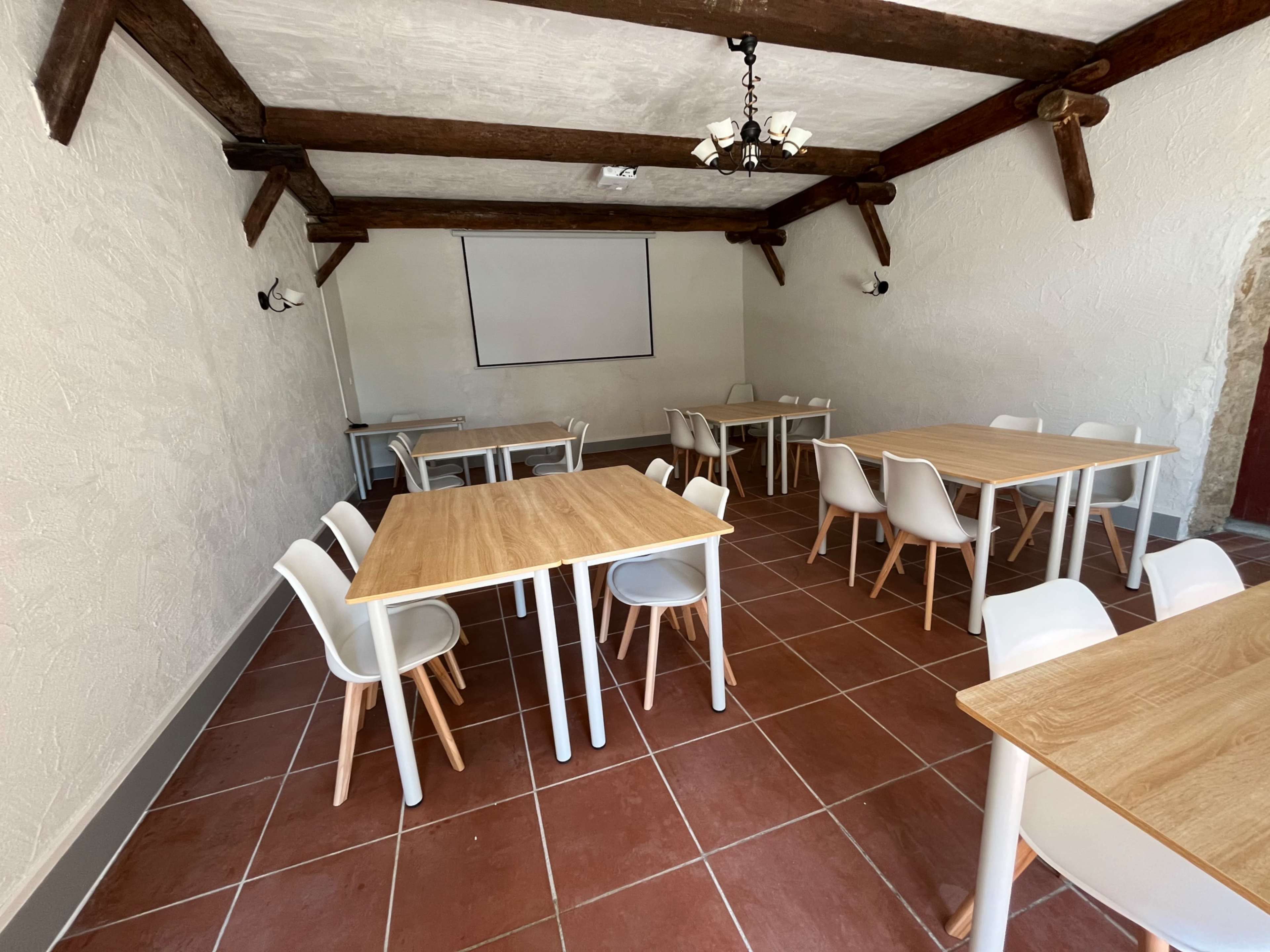 The image shows a simple, well-lit room with several tables and chairs arranged for a gathering or meeting, featuring wooden beams on the ceiling and tile flooring.