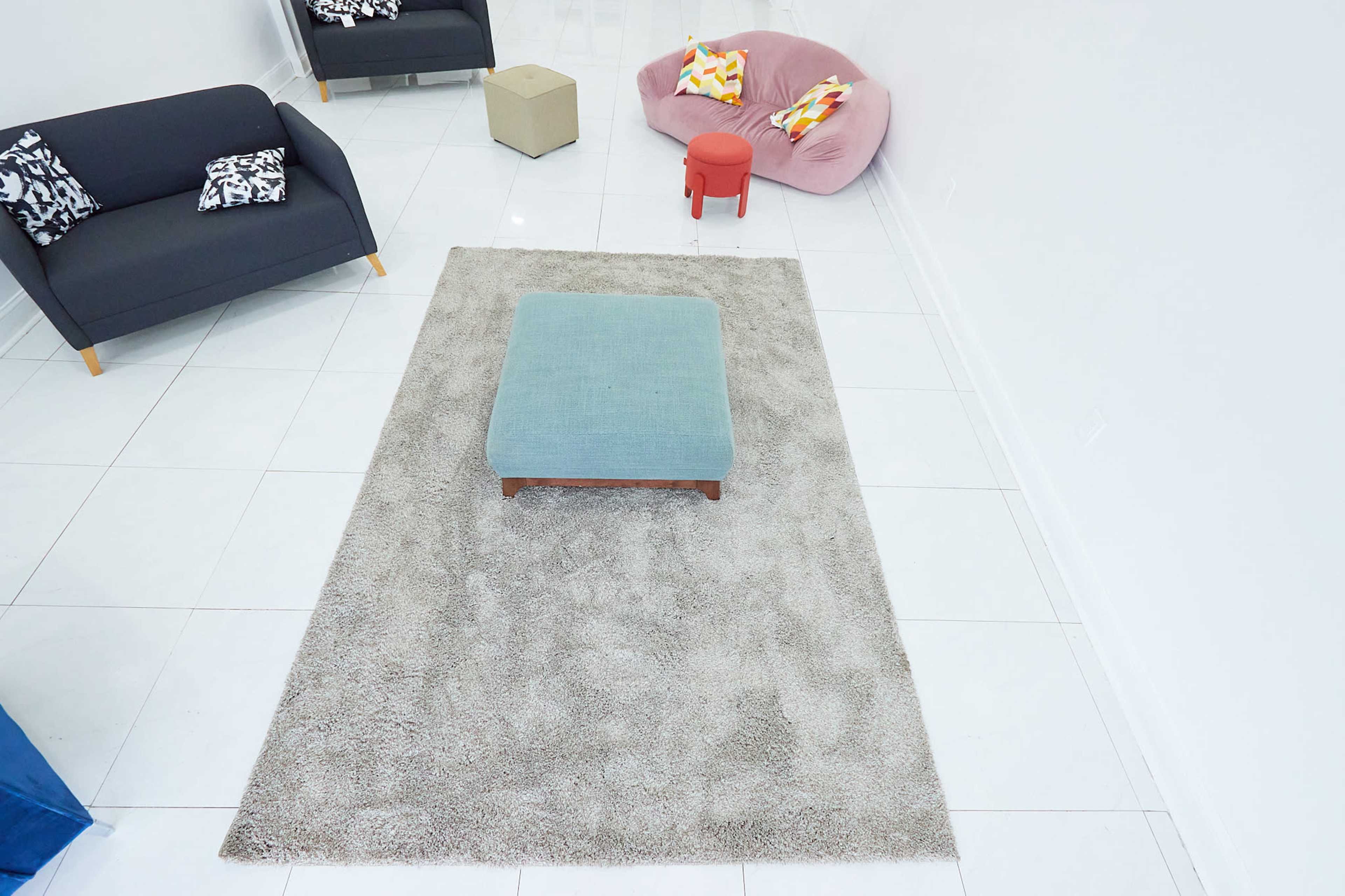 The image shows a modern, minimalist room with white tiled floors, a plush gray area rug, and various seating options, including a dark gray sofa, a pink beanbag chair, and a small red stool.