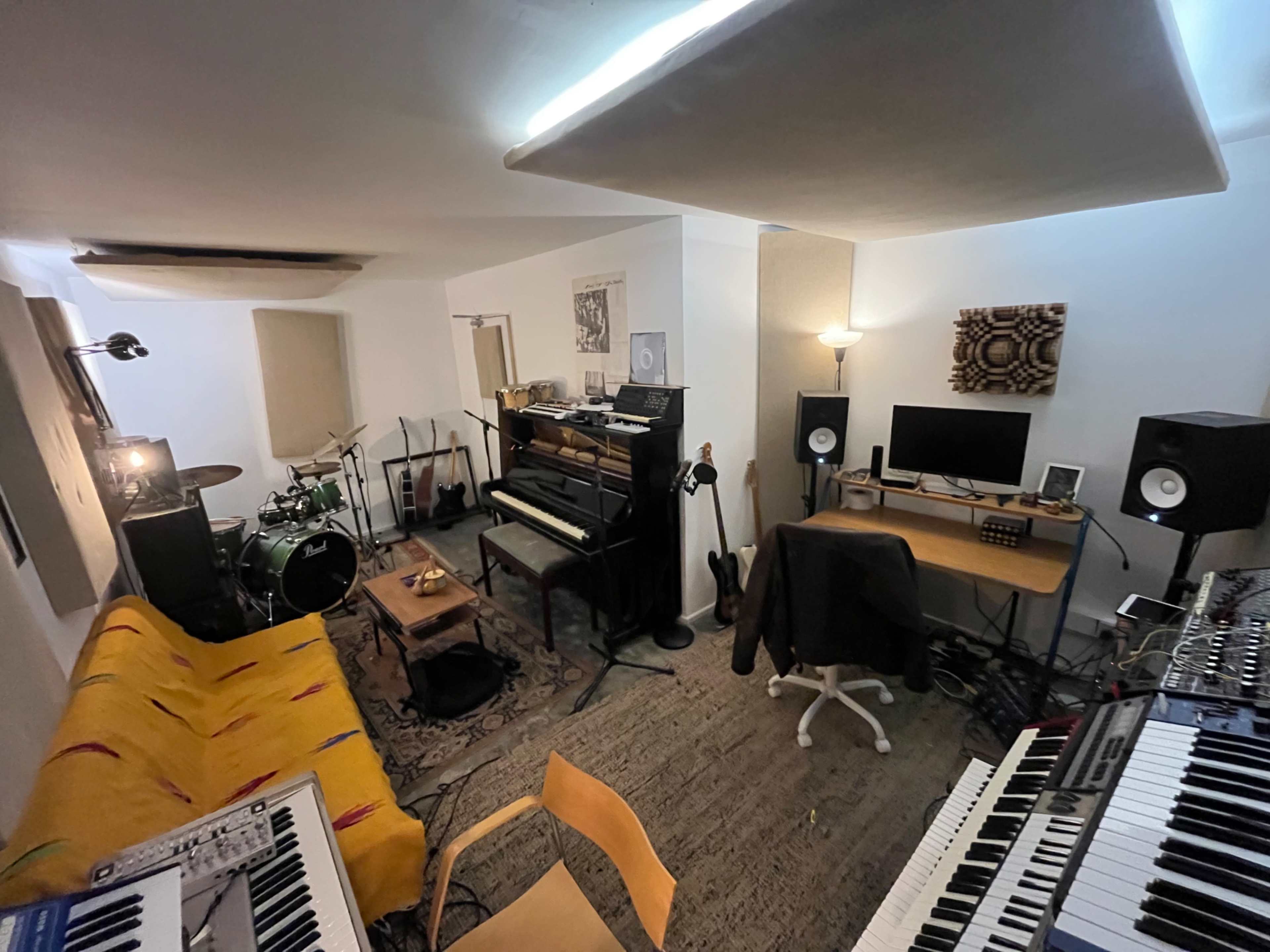 The image shows a music studio featuring various instruments including a drum set, piano, and synthesizers, with a desk and computer setup in the corner.