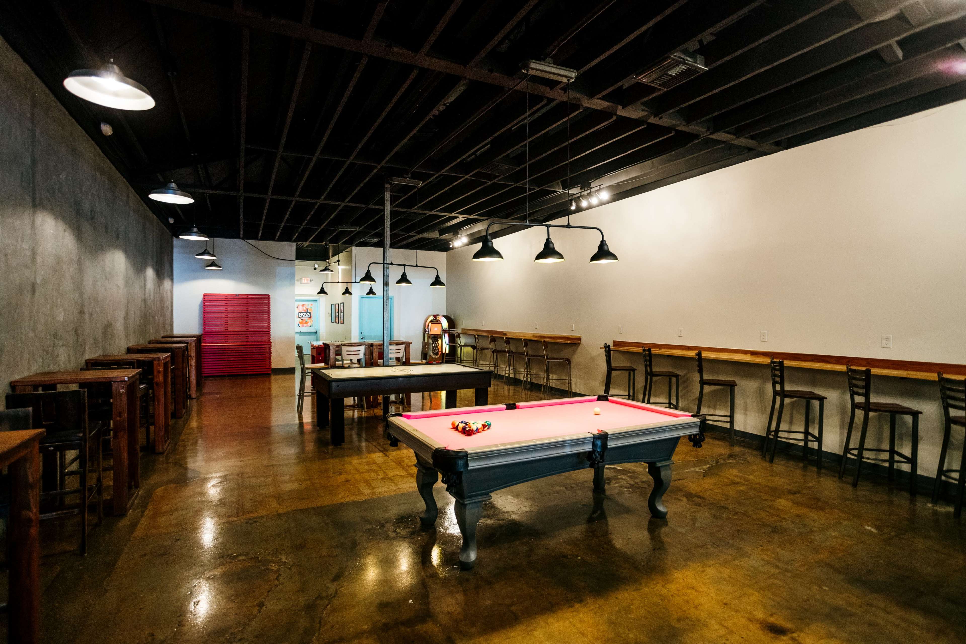A spacious game room features a pool table, a jukebox, and several tables with chairs arranged around a polished concrete floor.