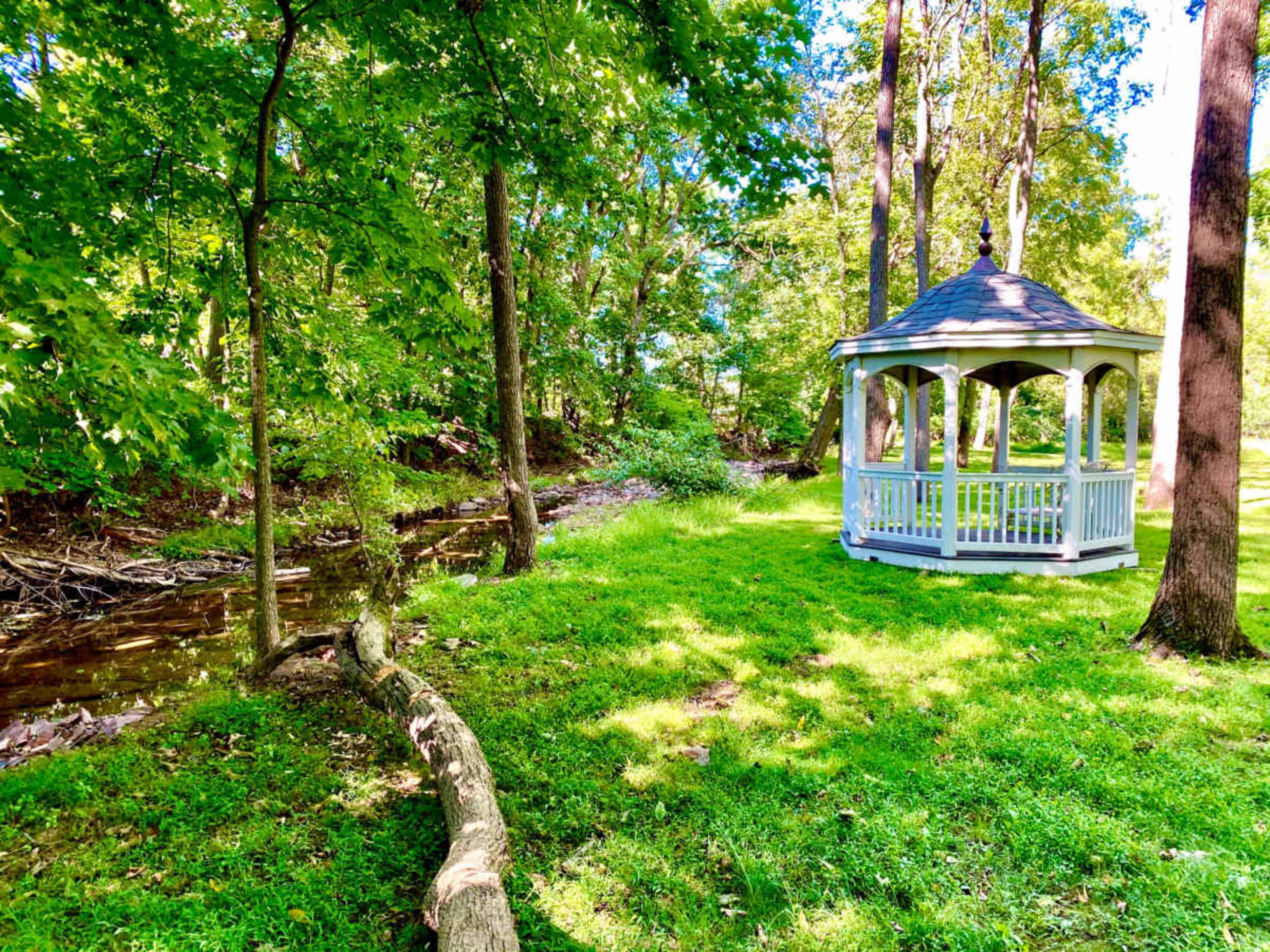 A white gazebo stands beside a small creek, surrounded by lush green grass and trees.