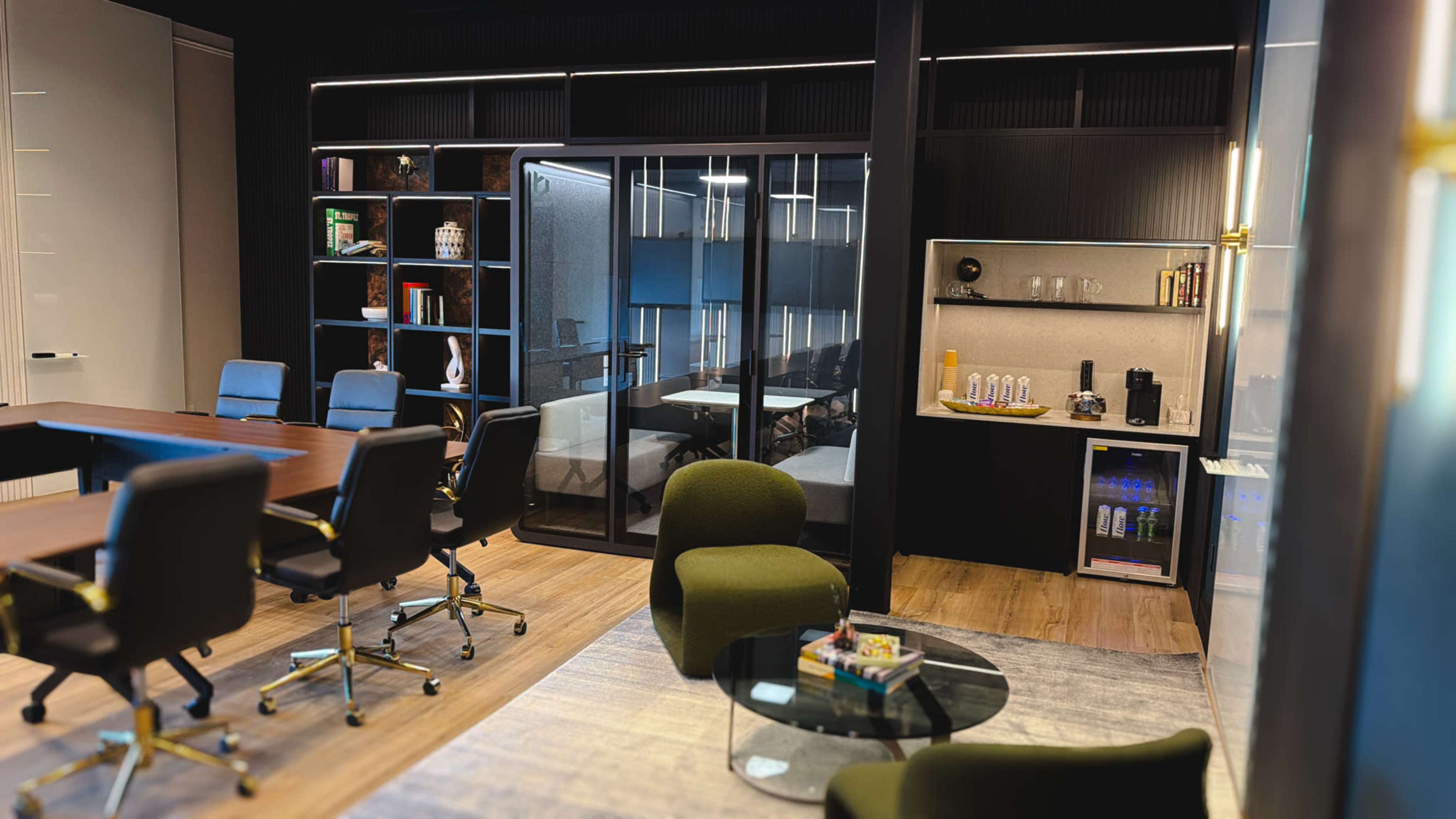 The image depicts a modern office space featuring a conference table, green seating, shelves with books, and a kitchenette area with a coffee machine and beverages.