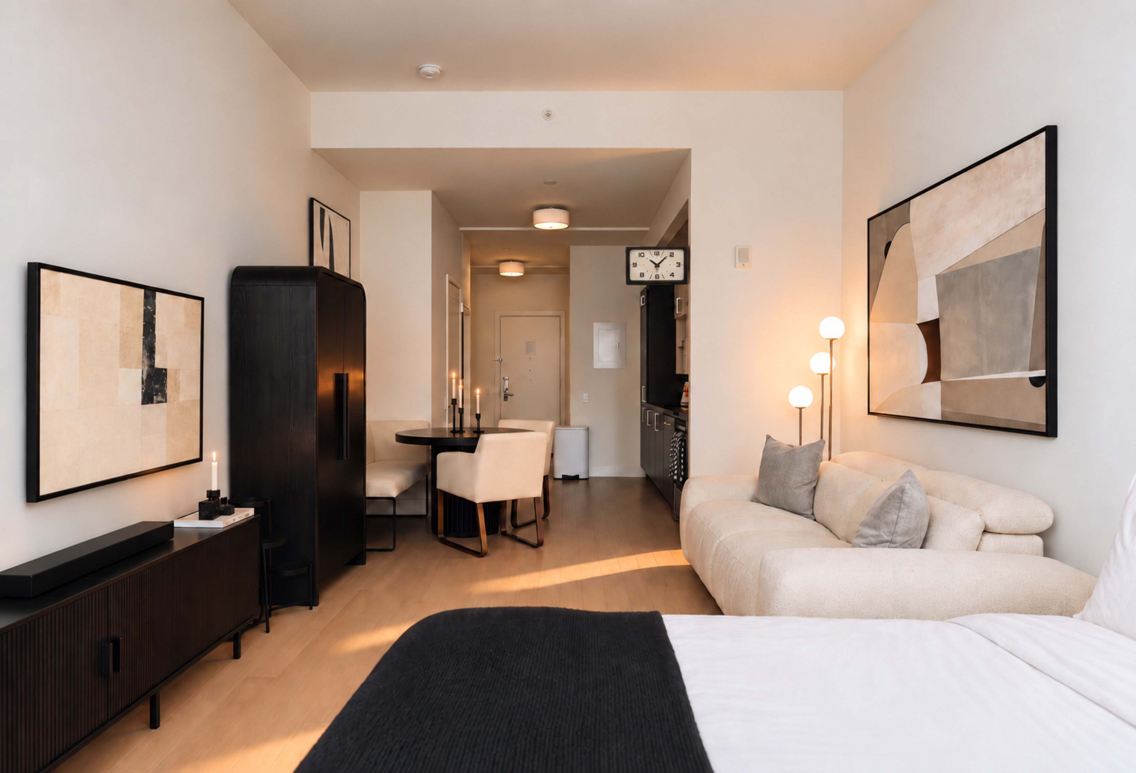 The image shows a modern studio apartment with a bed at the forefront, a seating area, a dining table, and kitchen appliances in the background.