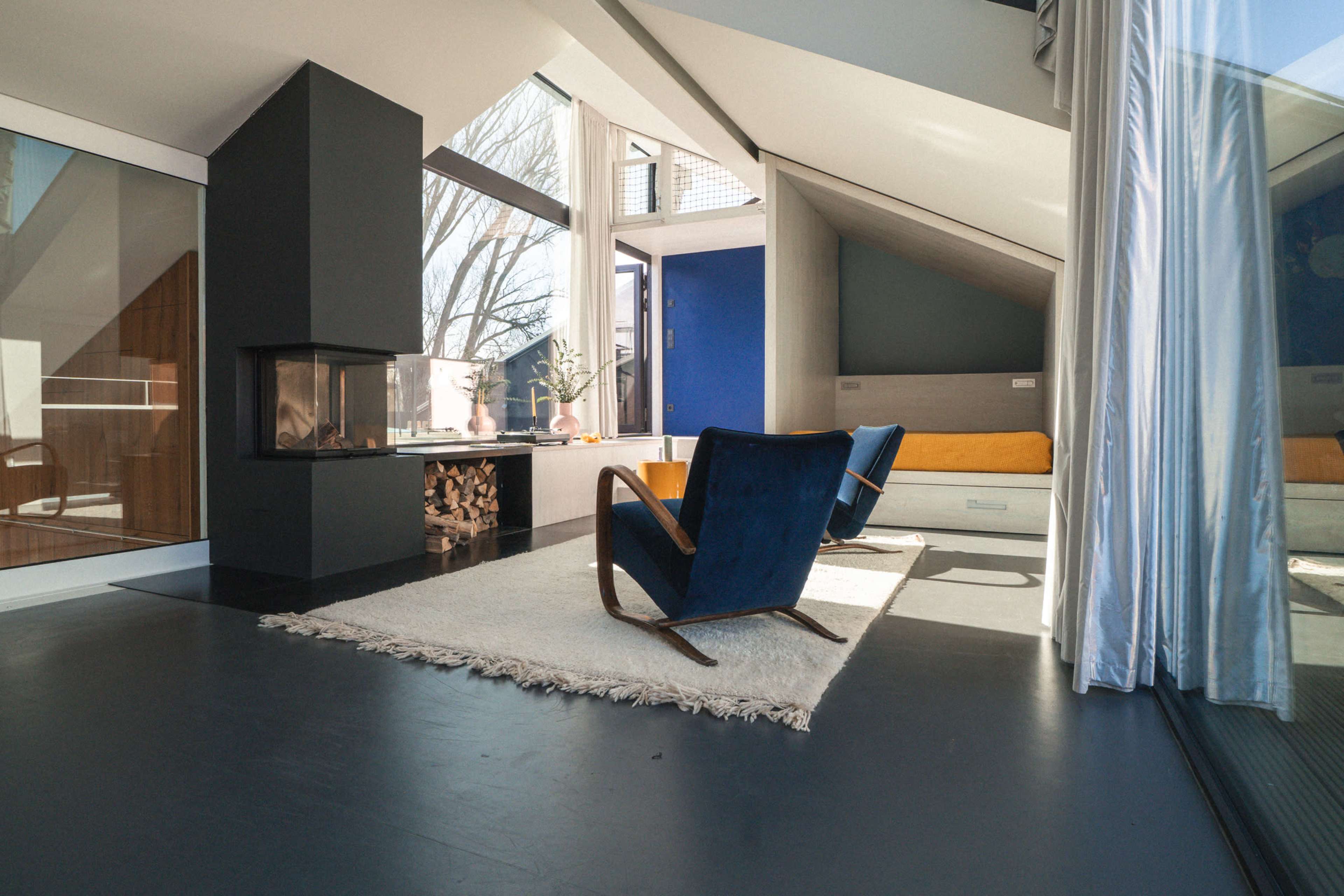 A modern living room features two blue chairs, a fireplace, large windows, and a light rug on a dark floor.