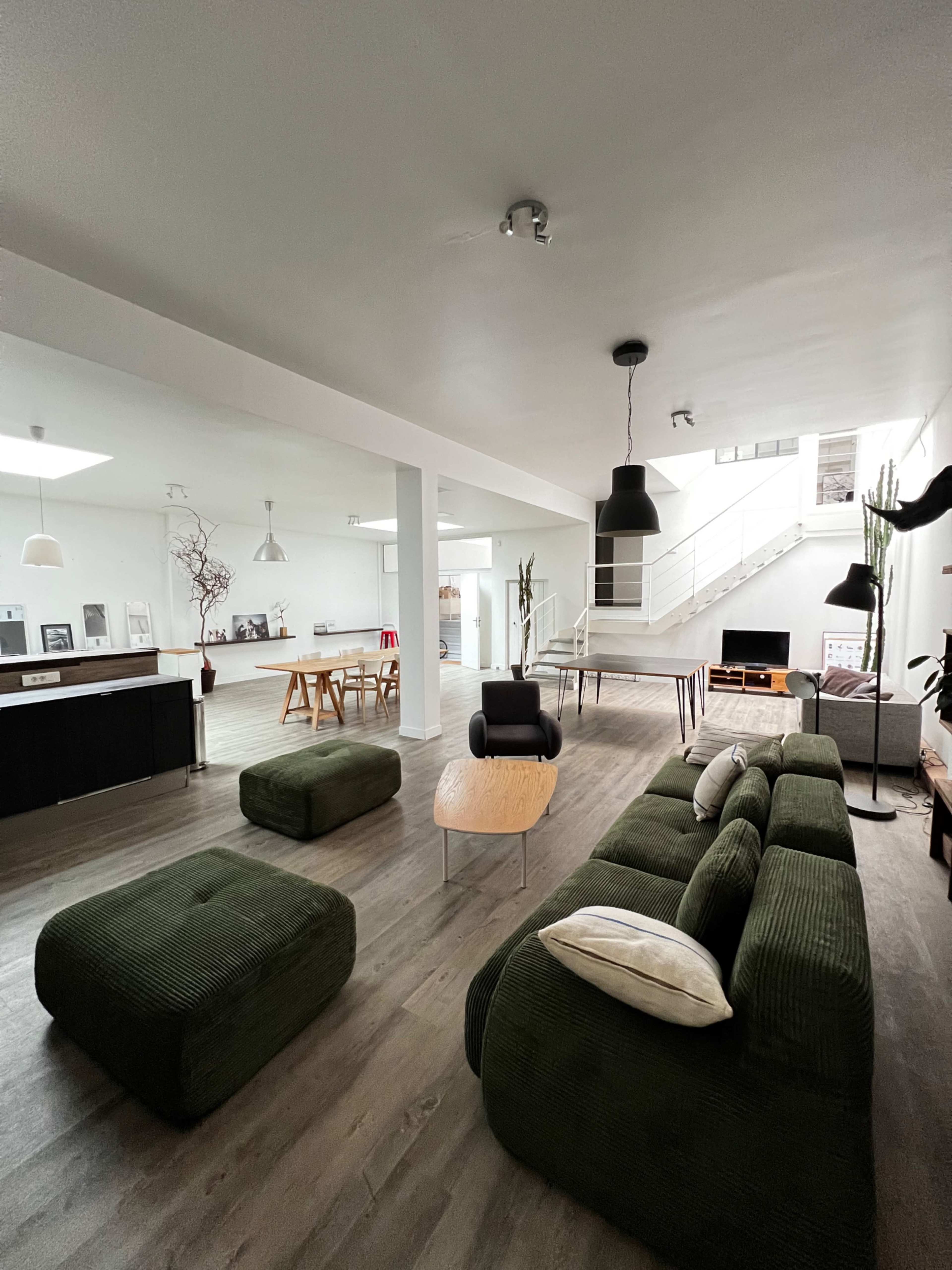 The image shows a modern, open-plan living space with green furniture, a wooden coffee table, and a dining area in the background.