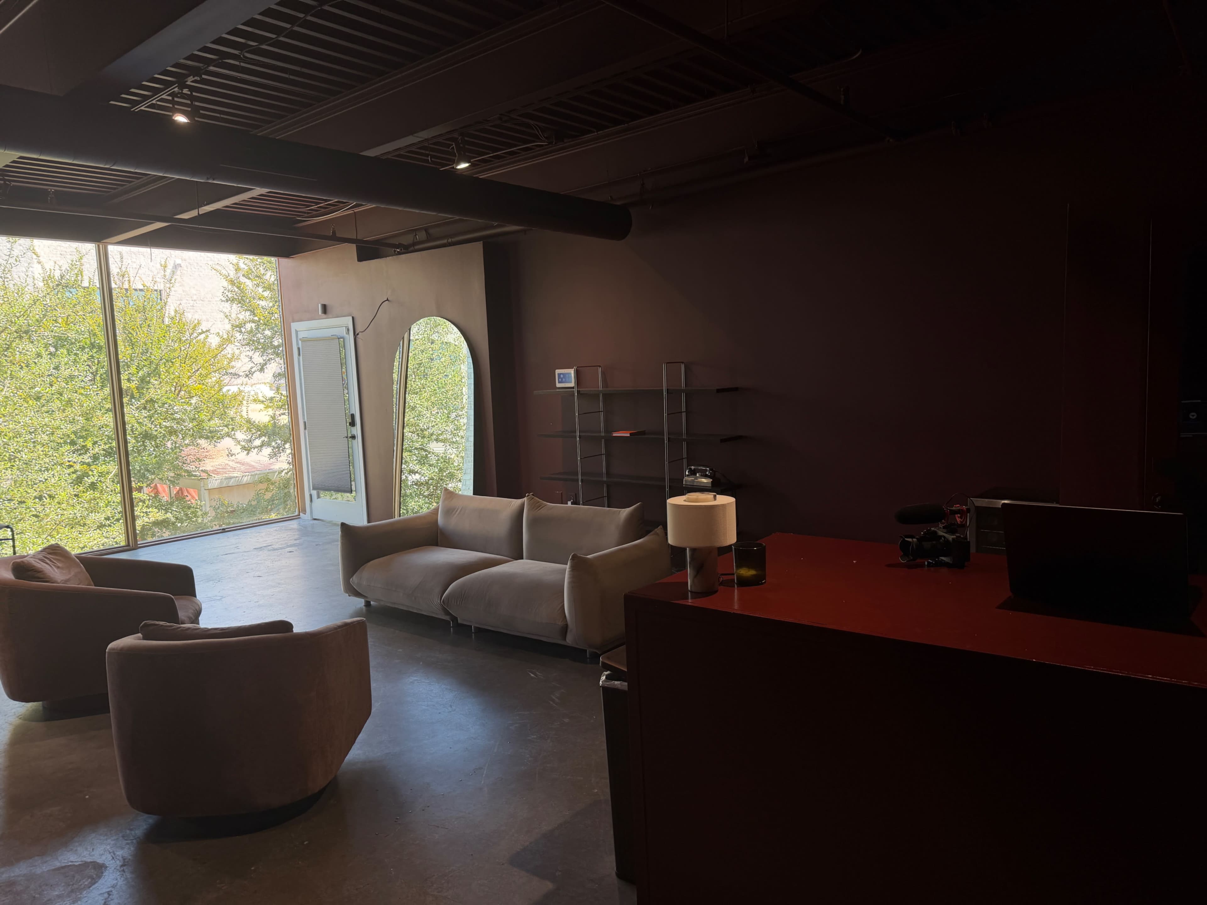The image shows a modern interior space featuring two armchairs, a couch, a lamp, and a large window with a view of greenery outside.