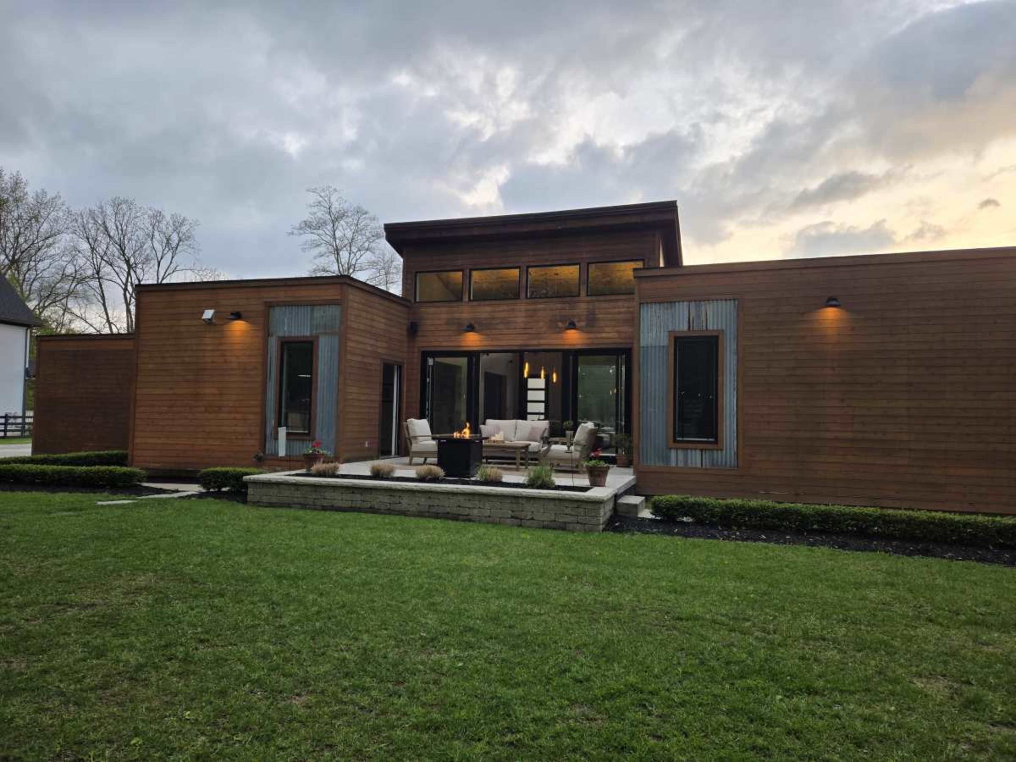 The image shows a modern wooden house with large glass windows and a stone patio surrounded by a green lawn.
