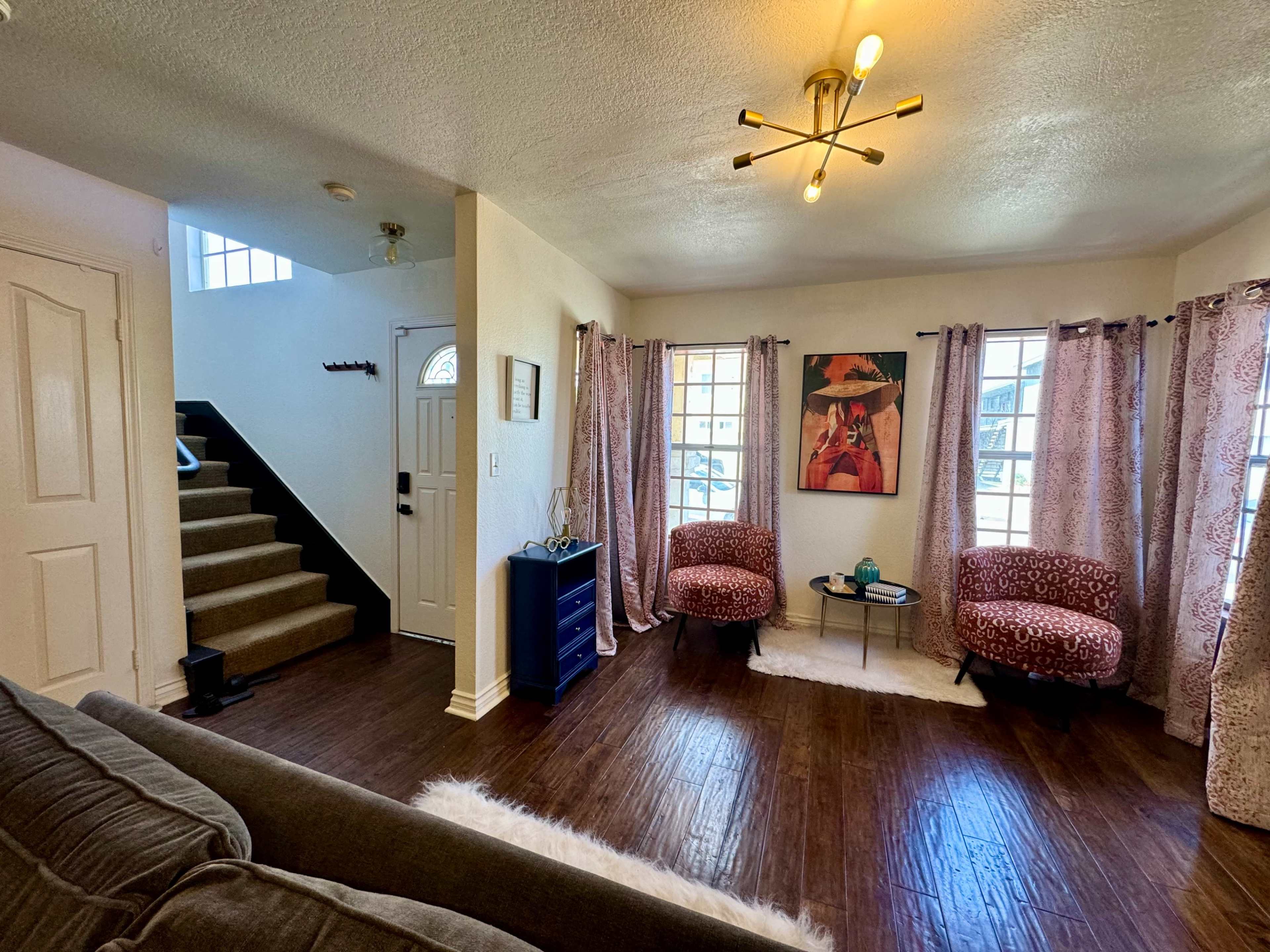 The image shows a well-lit living room with two patterned chairs, a small table, and a staircase leading to another level.