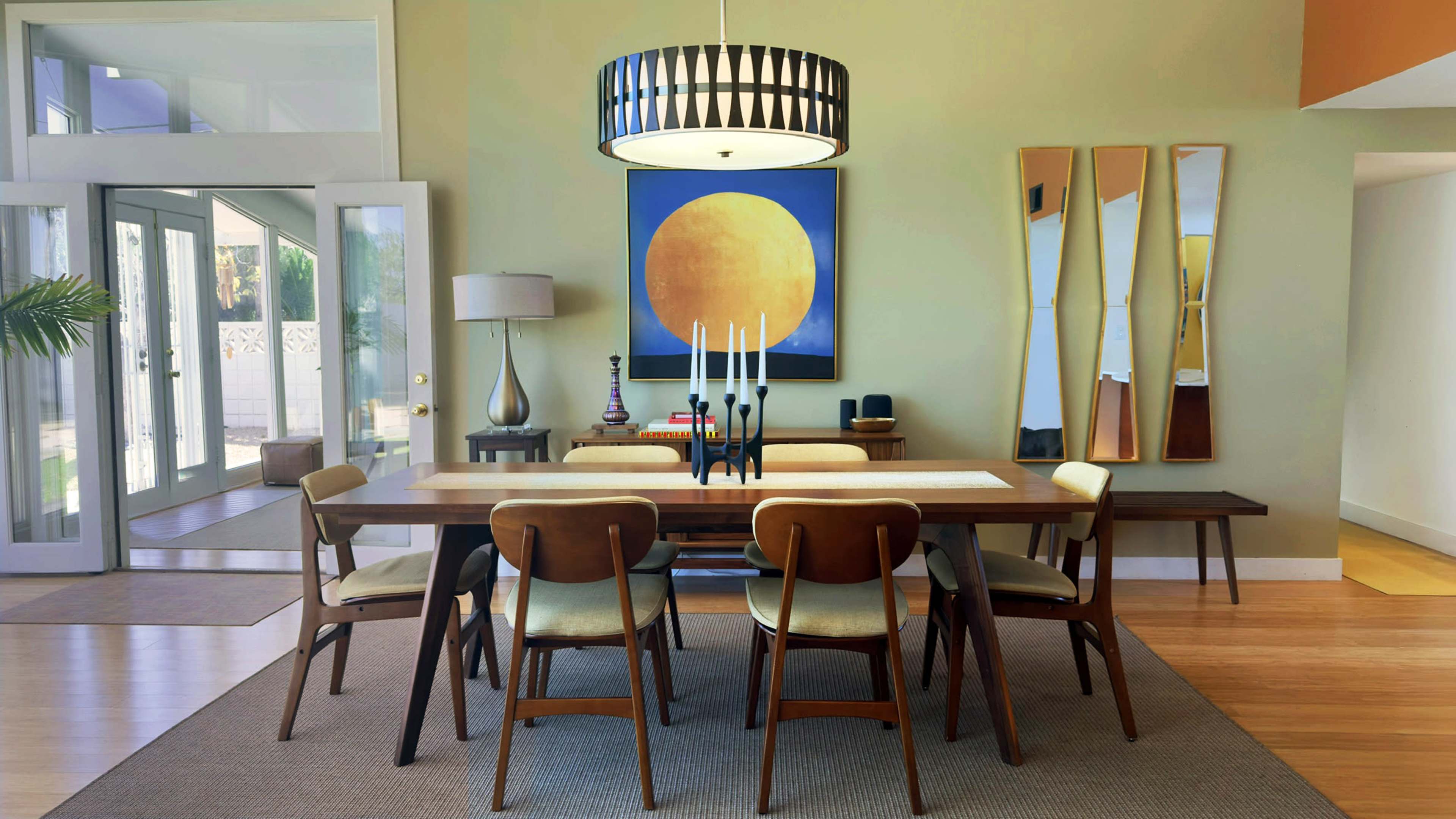 A modern dining room features a long wooden table surrounded by six chairs, with a large circular light fixture and a colorful abstract painting on the wall.