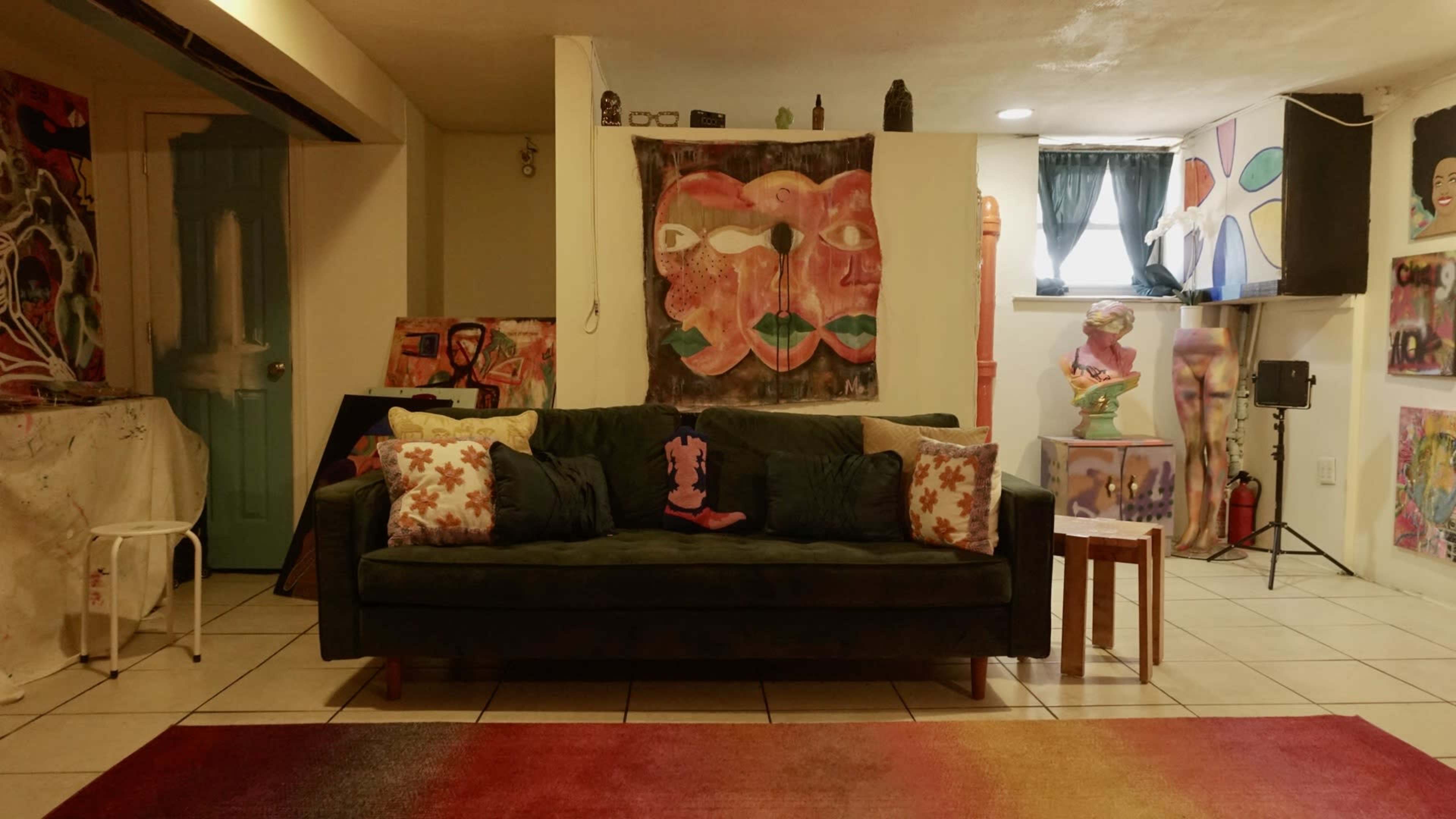 A cozy room featuring a green couch with decorative pillows, surrounded by various pieces of colorful artwork on the walls.