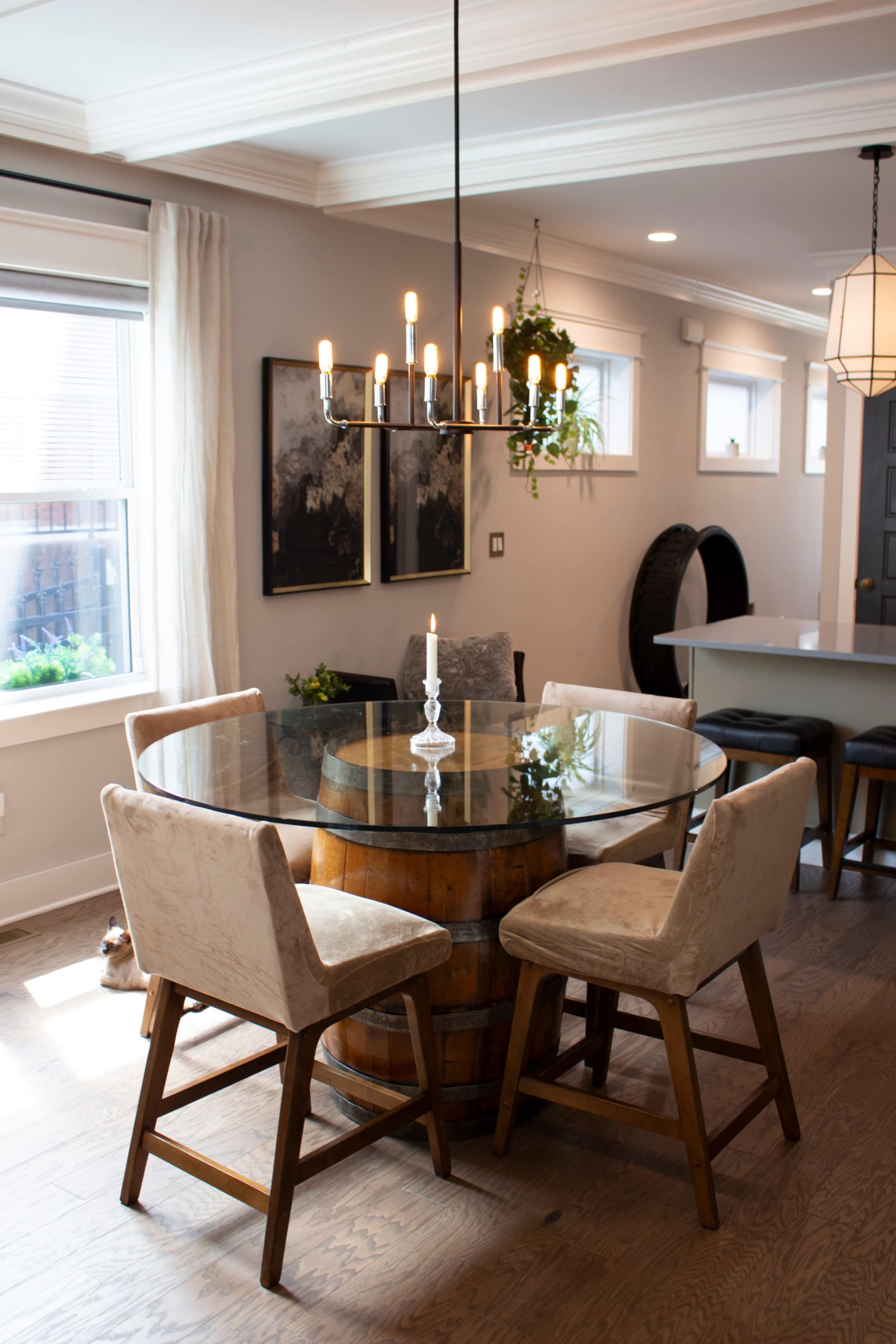 The image shows a modern dining area featuring a circular glass table atop a wooden barrel, flanked by two upholstered chairs and illuminated by a chandelier.