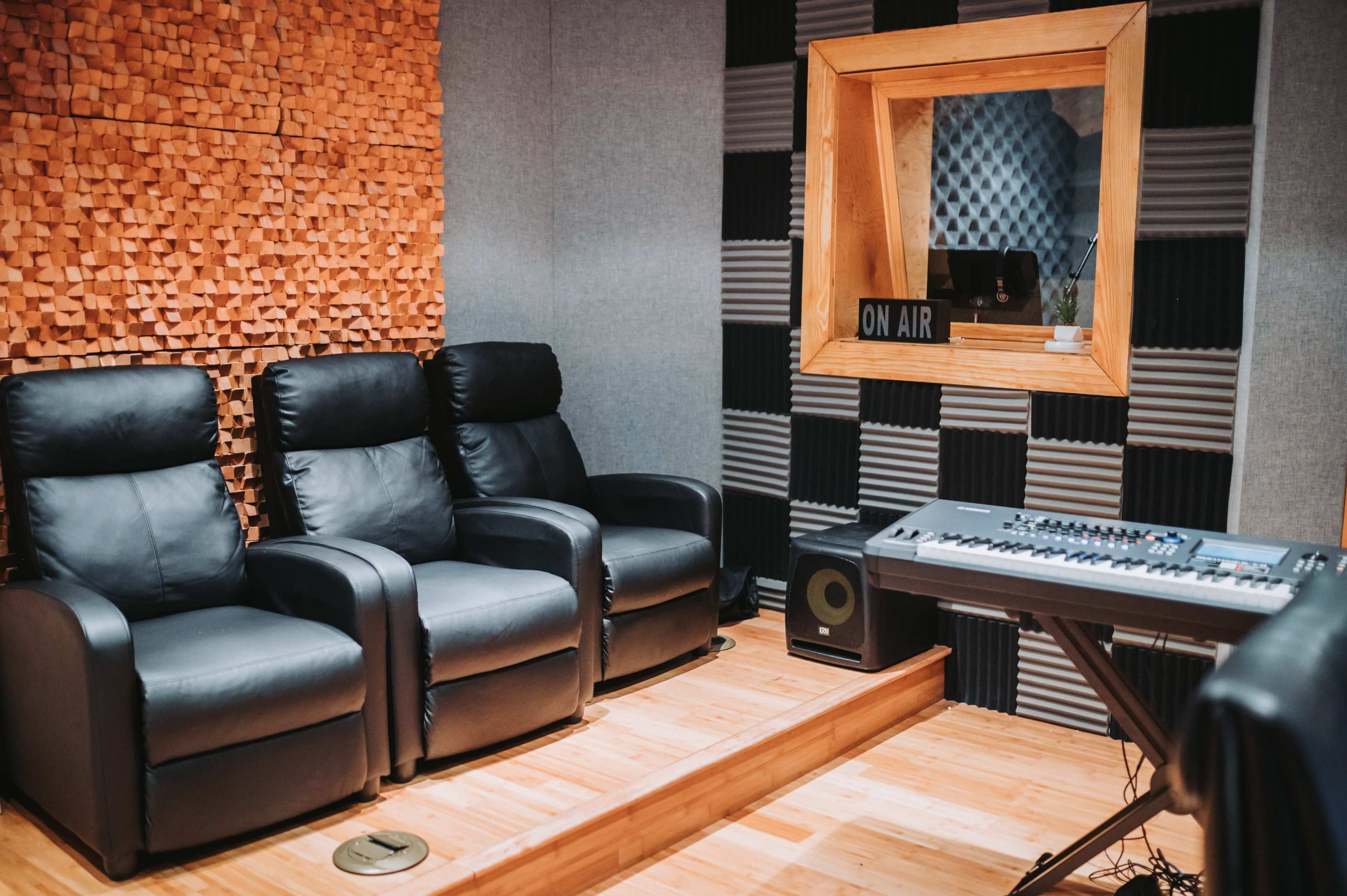 The image shows a sound studio featuring three black reclining chairs, a keyboard, and a soundproof wall with textured panels.