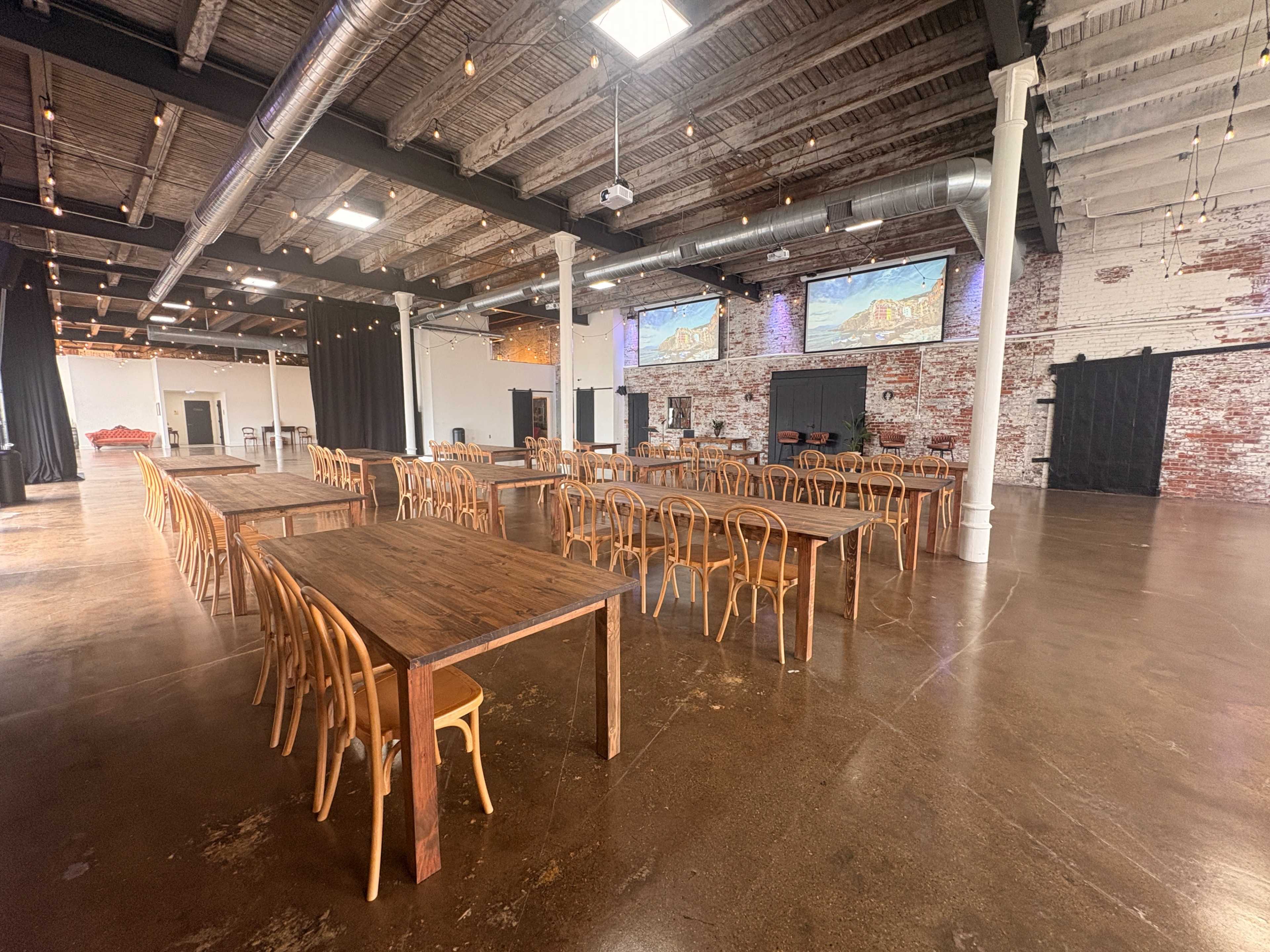 The image shows a large, industrial-style event space with wooden tables arranged in rows, surrounded by exposed brick walls and lit by overhead string lights.