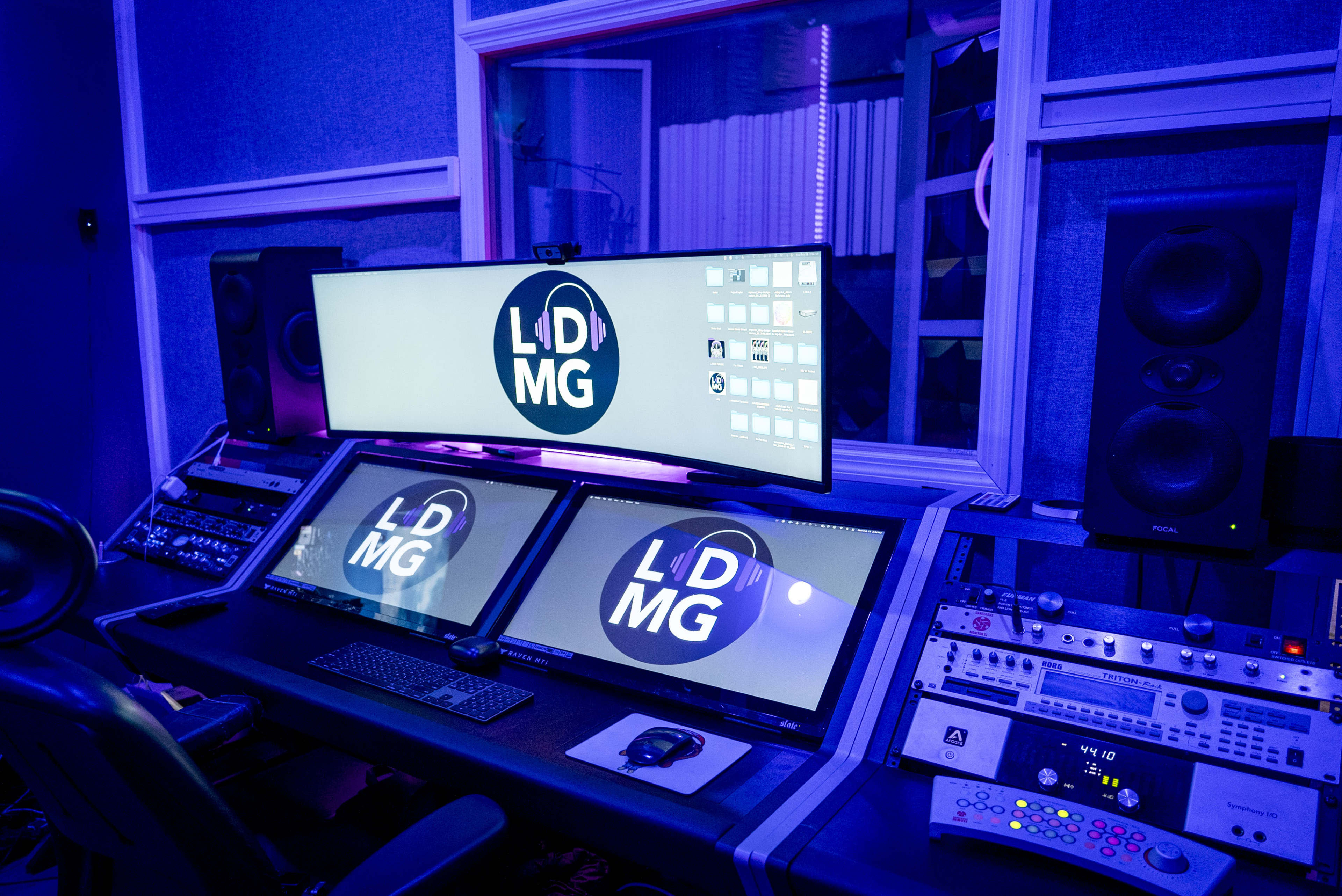 A music studio with two large monitors displaying a logo, mixing equipment, and speakers, all illuminated by blue lighting.