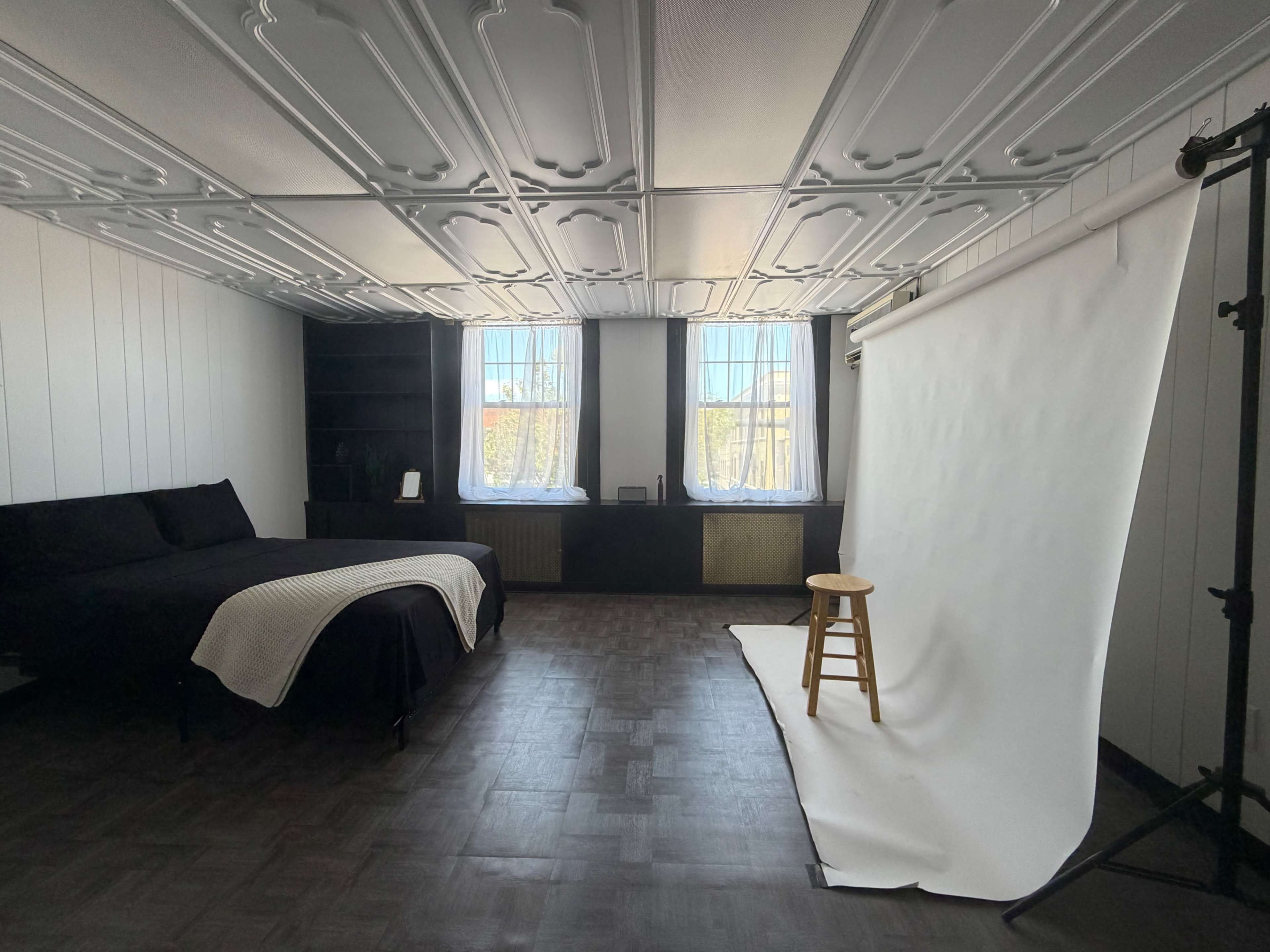 The image shows a spacious room with a black bed, a white backdrop on a stand, and large windows letting in natural light.