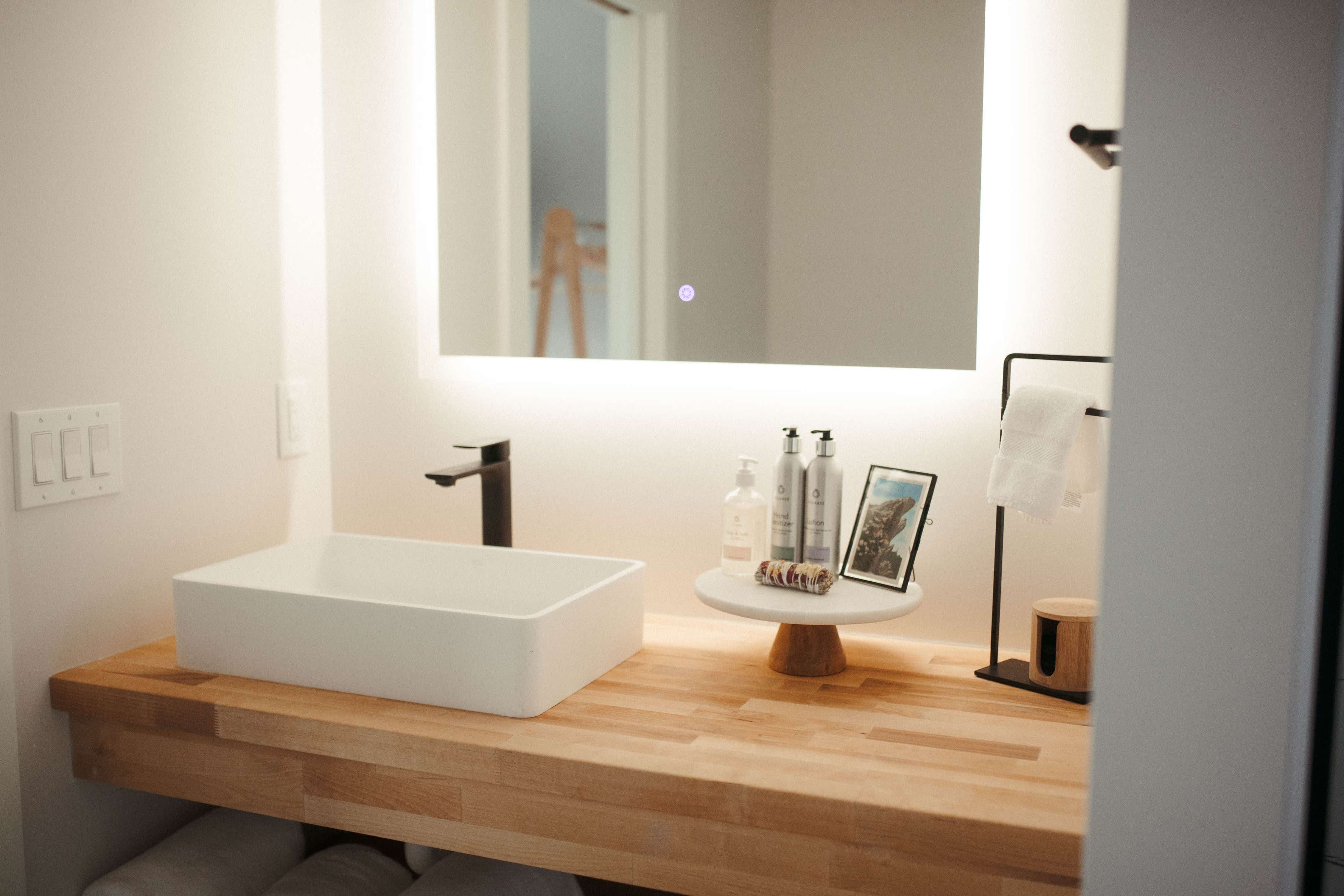 A modern bathroom features a wooden countertop with a rectangular sink, a round display stand for toiletries, and a backlit mirror.