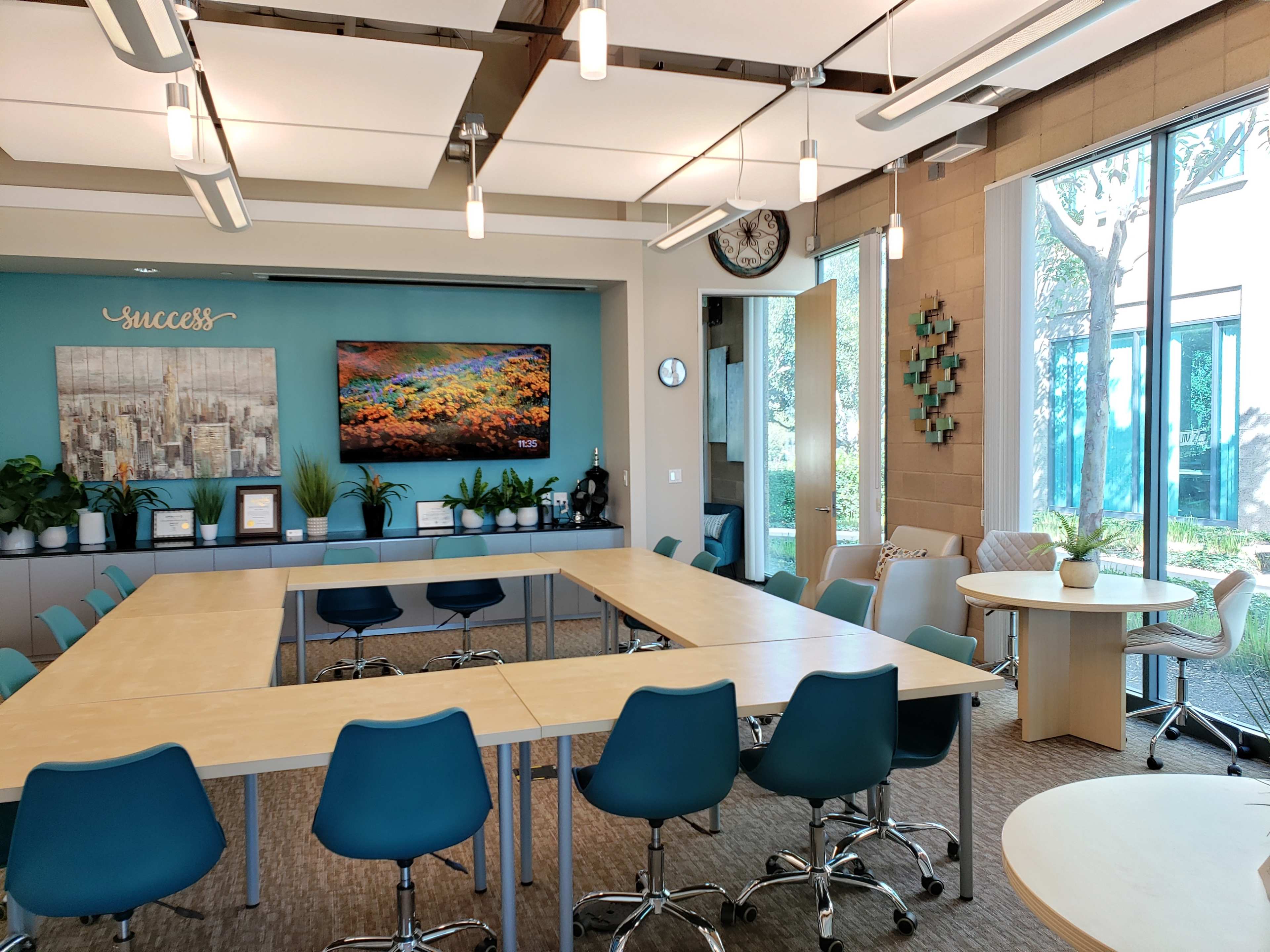 A modern conference room with multiple tables arranged in a U-shape, surrounded by teal chairs, and features large windows with plants and artwork on the walls.