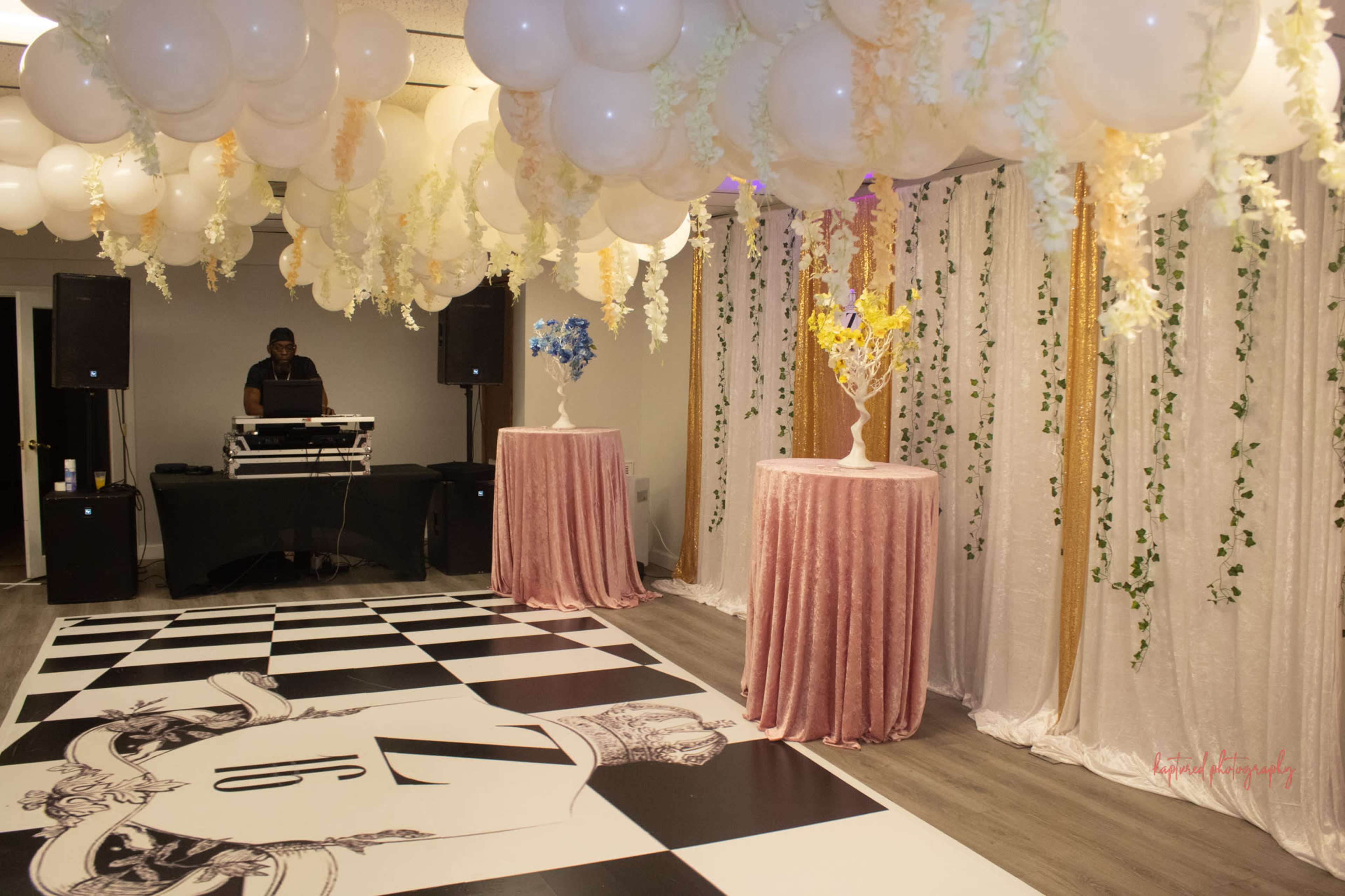 The room features a checkered dance floor with two circular tables adorned with floral arrangements and a DJ setup in the background, all beneath a canopy of balloons and draping.