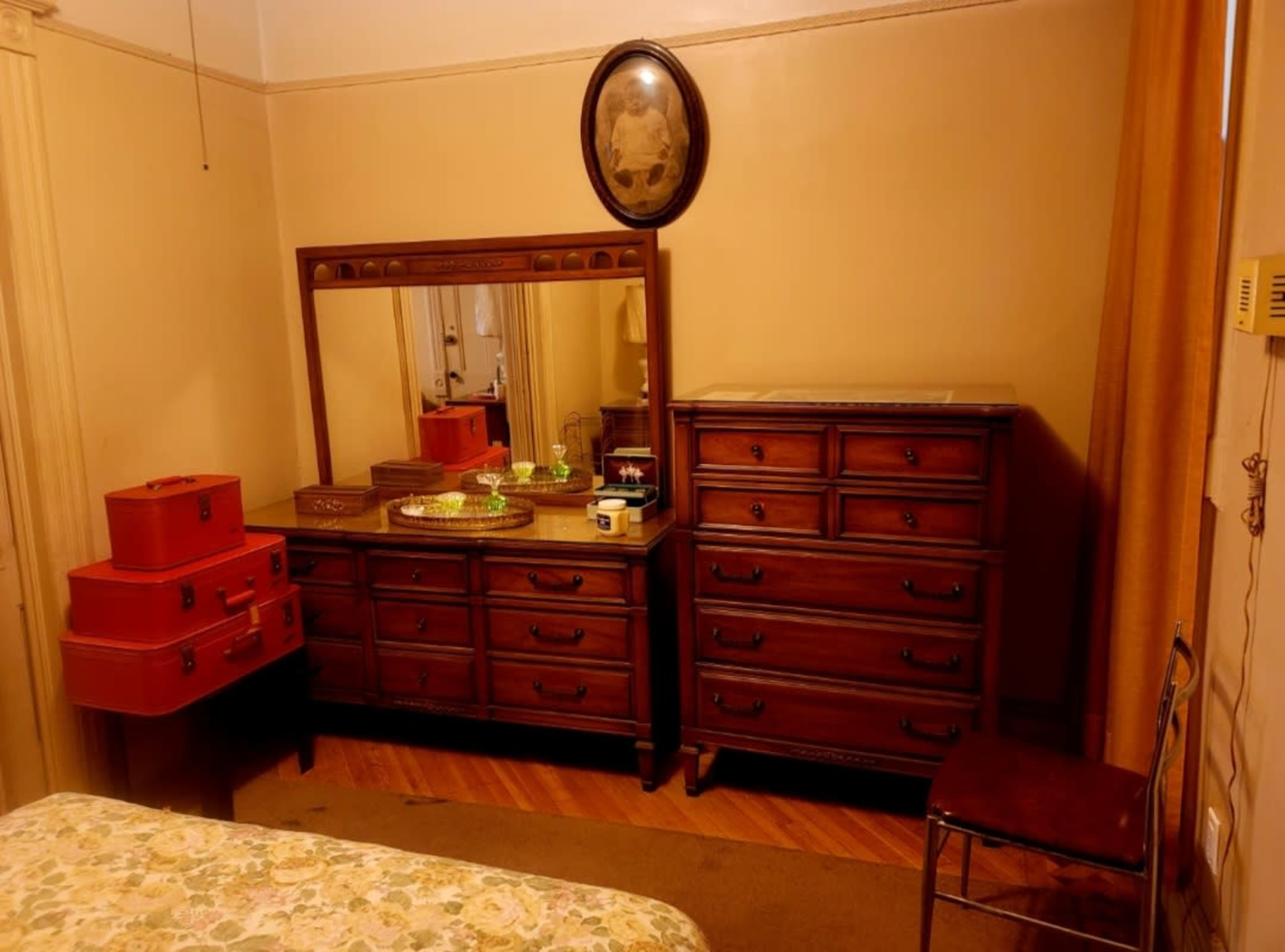 The image shows a vintage bedroom with a wooden dresser and mirror, a matching chest of drawers, and red storage boxes, all set against a yellow-walled backdrop.