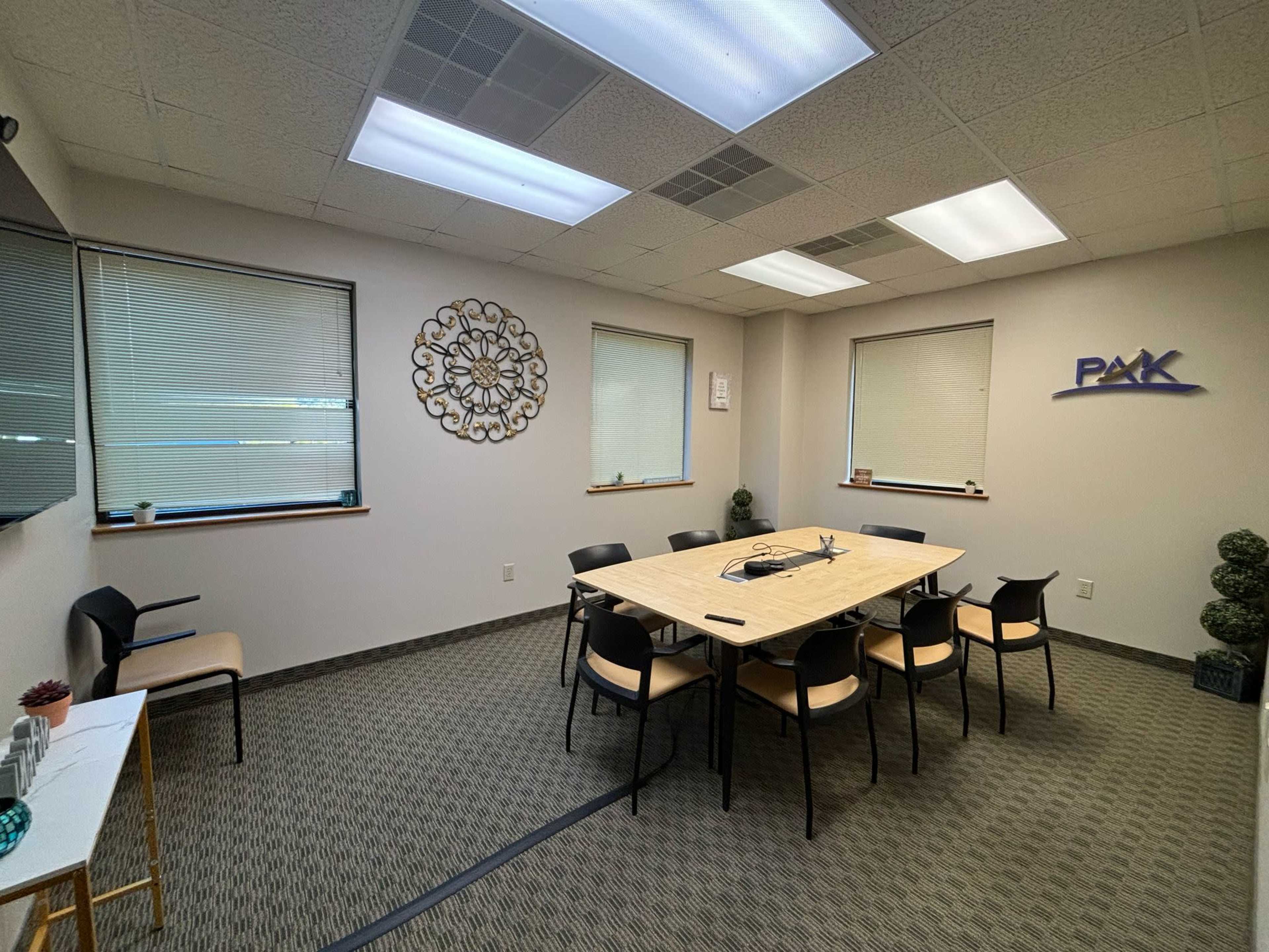 A conference room features a large, rectangular table surrounded by black chairs, with windows providing natural light and decor on the walls.