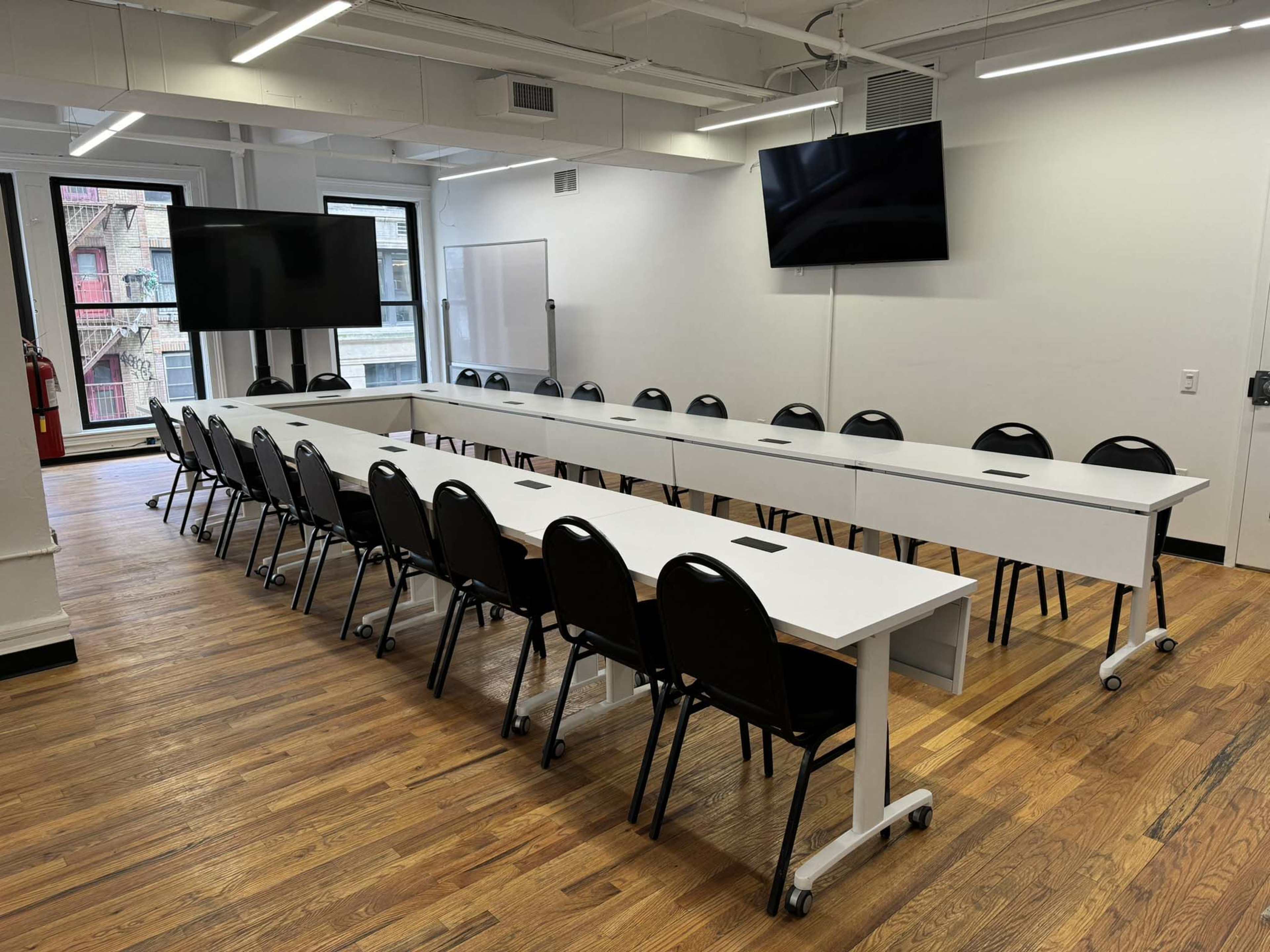 A conference room features two long tables arranged in a U-shape with black chairs and two wall-mounted screens.