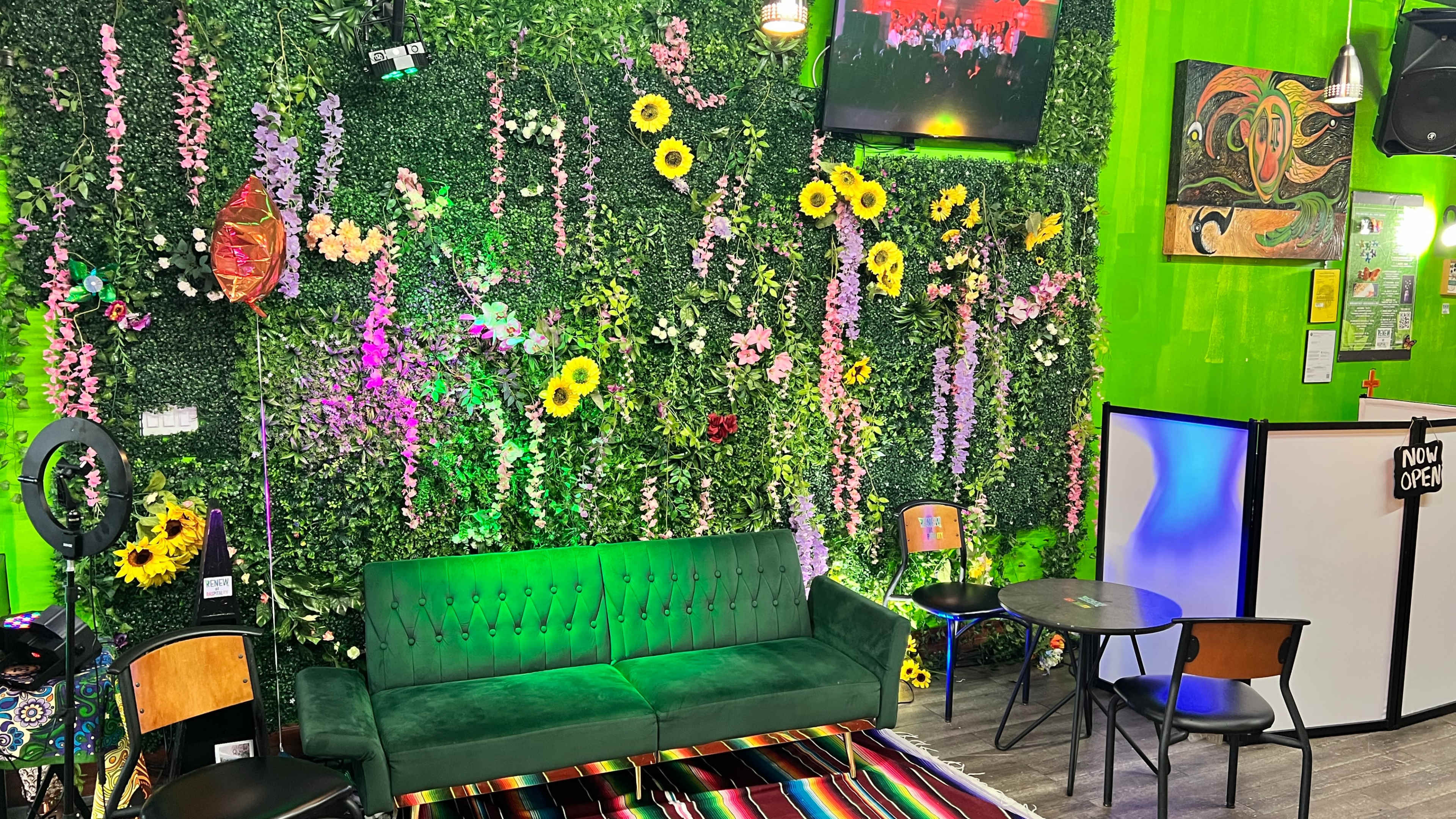 The interior of a vibrant café features a green wall adorned with artificial flowers, a green velvet couch, and colorful decorations.