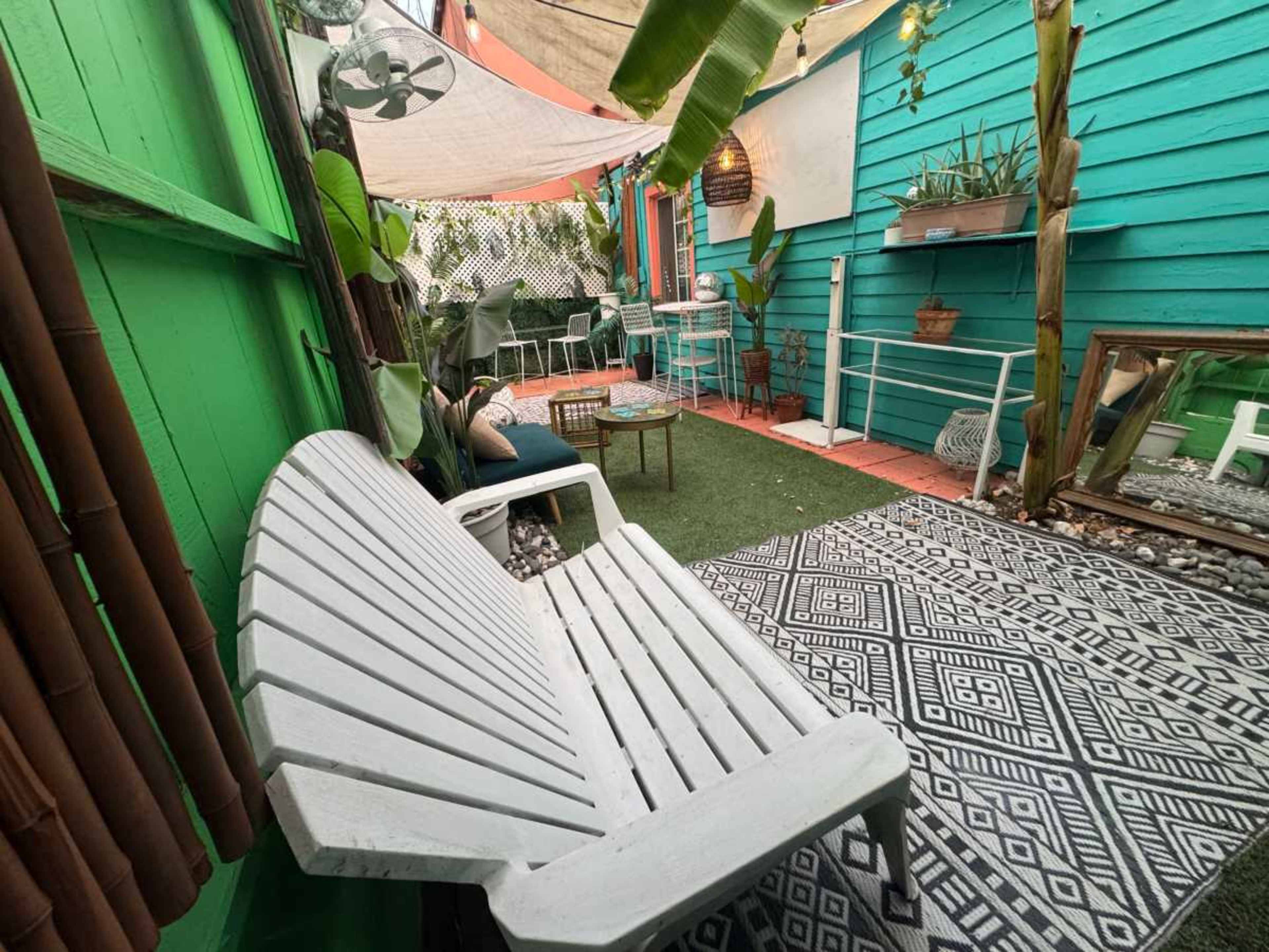 The image shows a colorful outdoor patio area featuring a white lounge chair, green walls, potted plants, and a patterned rug.