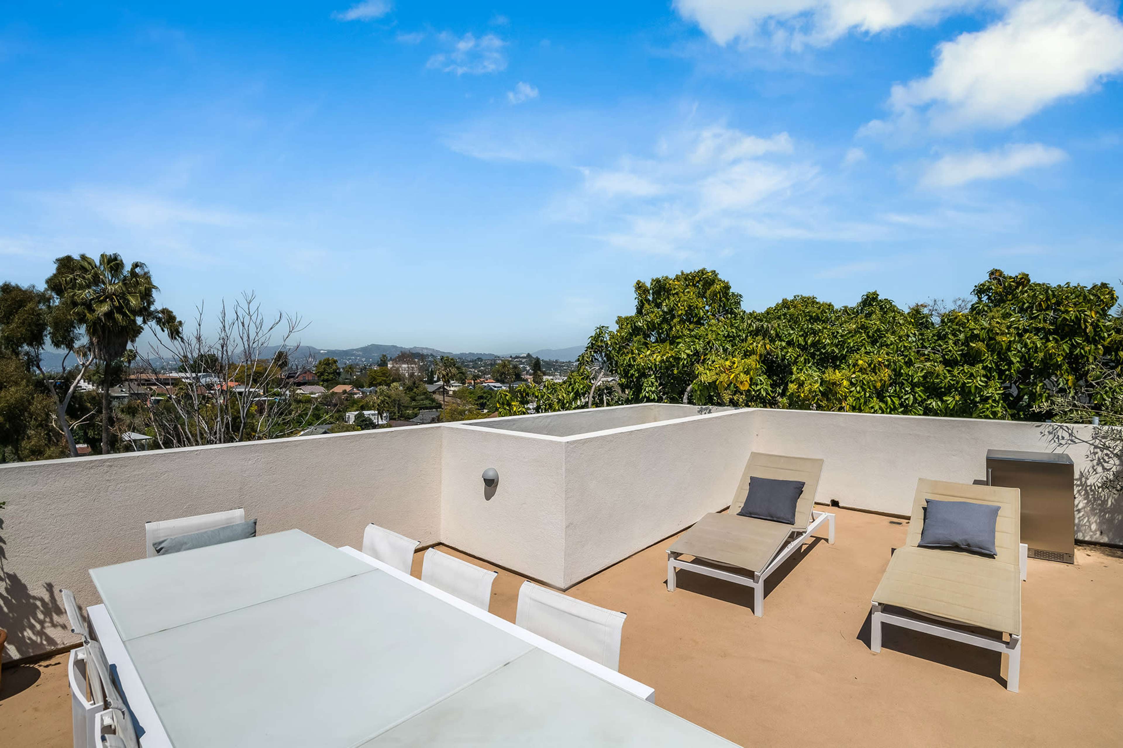 The image shows a rooftop patio with two lounge chairs, a dining table, and views of surrounding trees and distant hills under a blue sky.