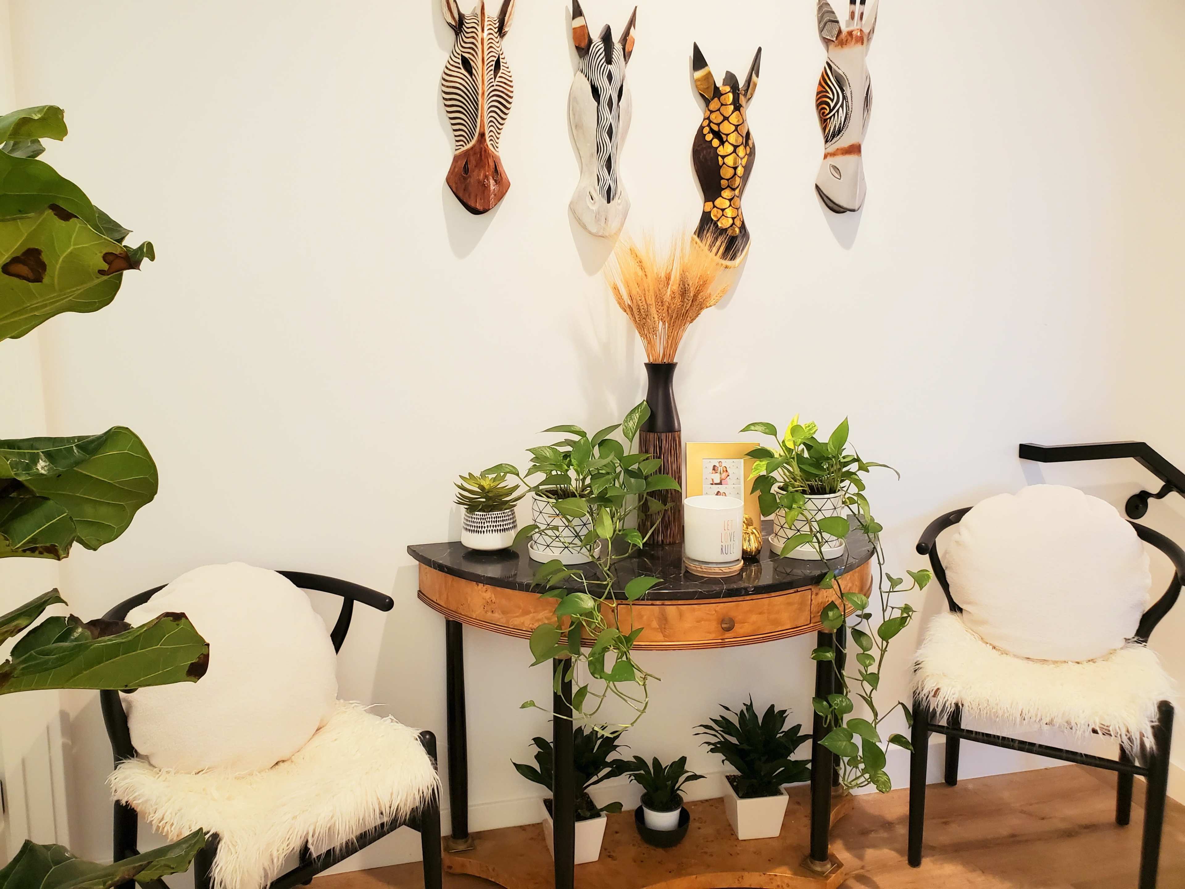 A small entryway features a round table adorned with plants and a decorative vase, flanked by two upholstered chairs, with art masks mounted on the wall above.