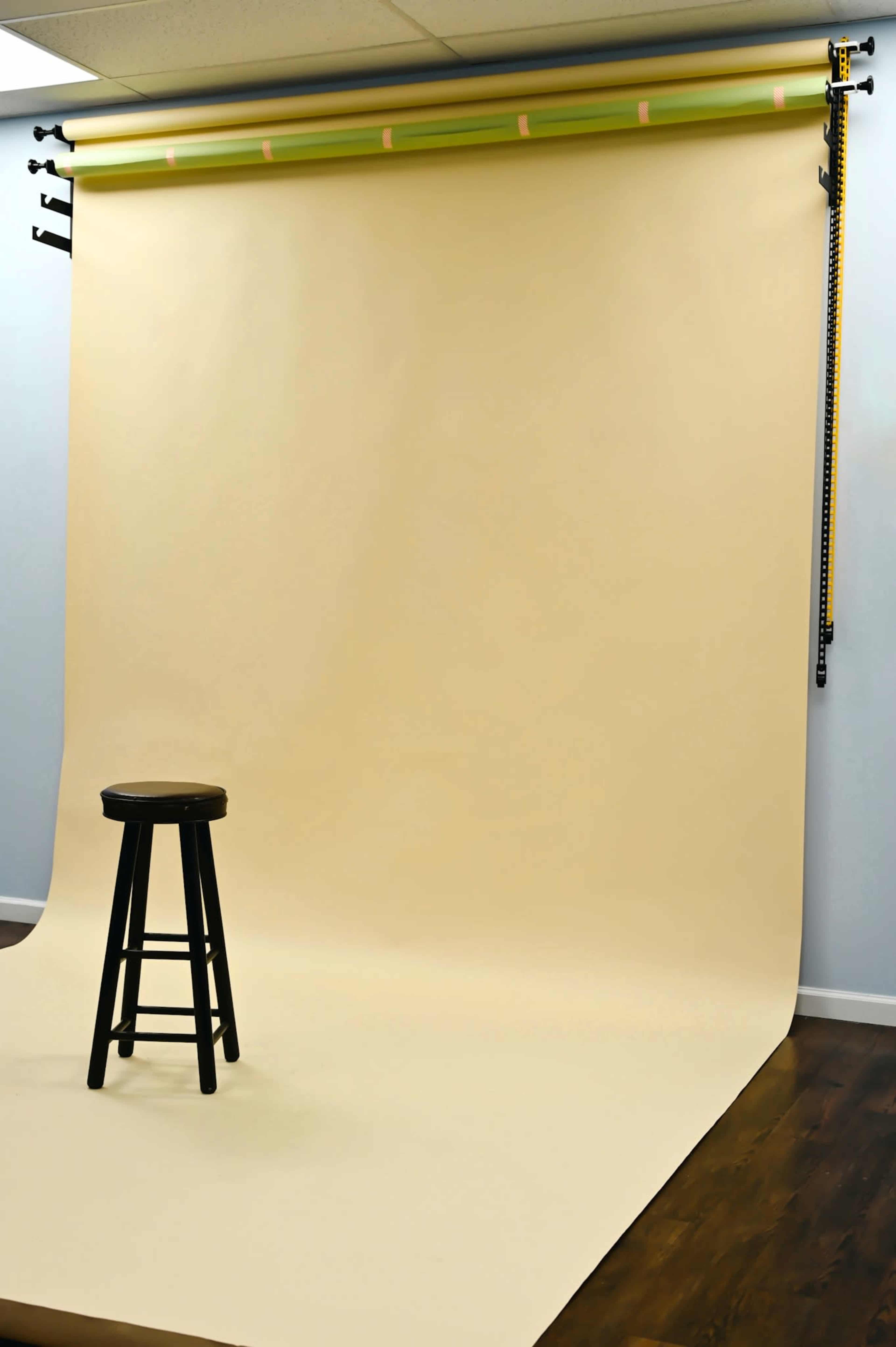 The image shows a photography studio with a large, beige backdrop and a black stool positioned on a wooden floor.