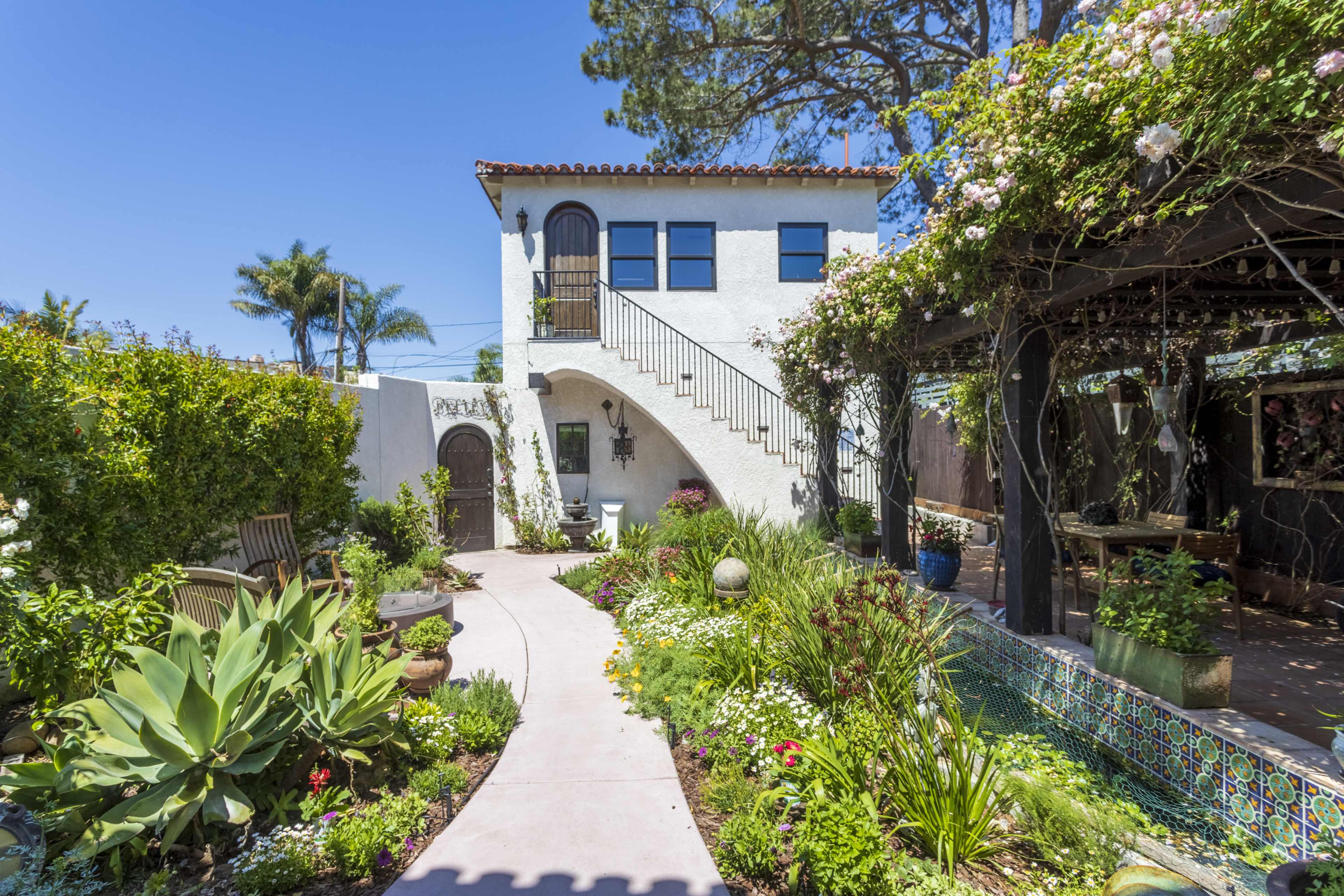 A pathway leads through a garden filled with various plants and flowers to a two-story white house with a staircase and a covered seating area adorned with greenery.