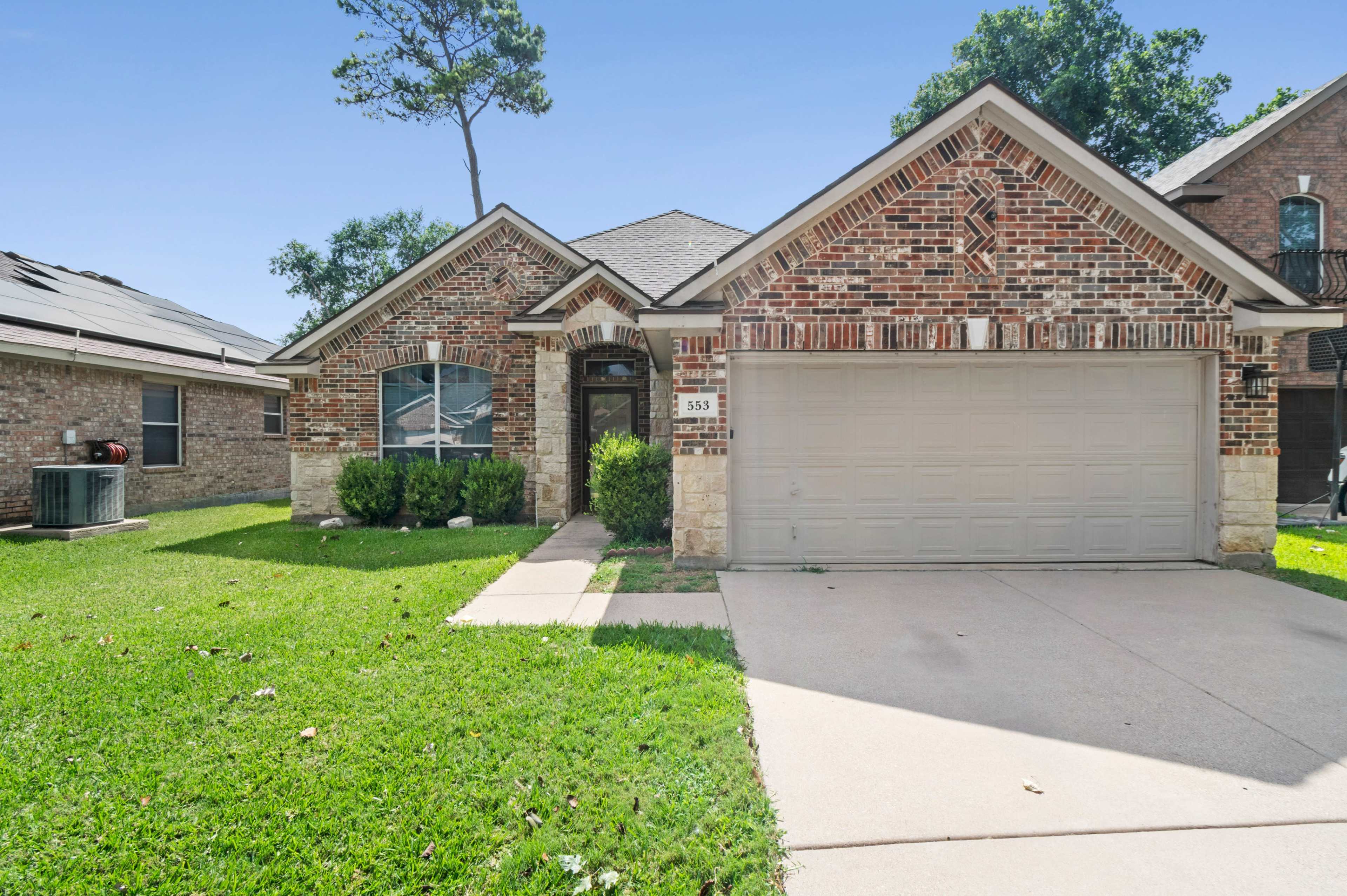 The image shows a single-story brick house with a two-car garage, a front yard with green grass, and a clear blue sky.