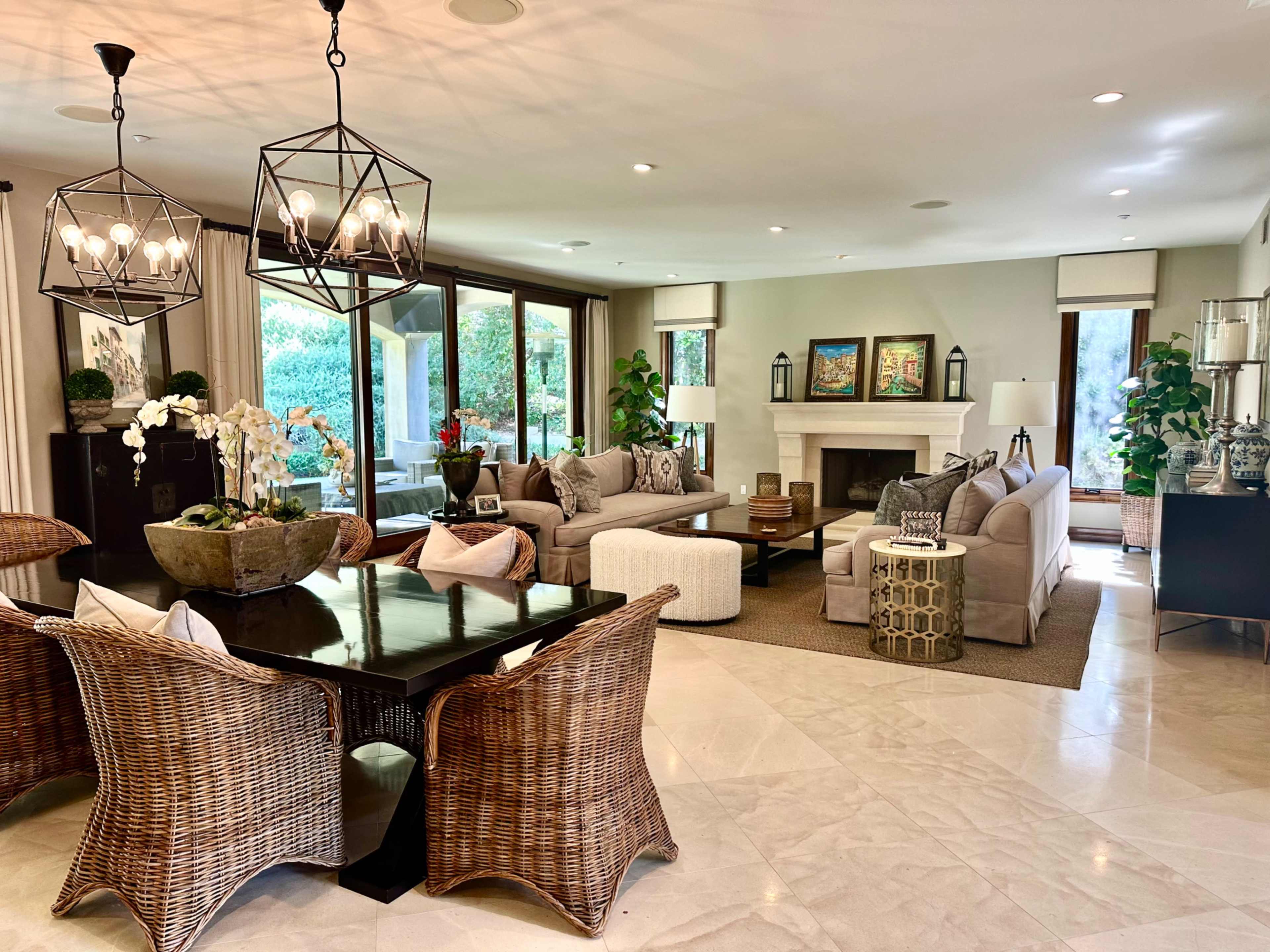 The image shows a spacious living room with a dining area, featuring a large table surrounded by wicker chairs and a comfortable seating arrangement near a fireplace.