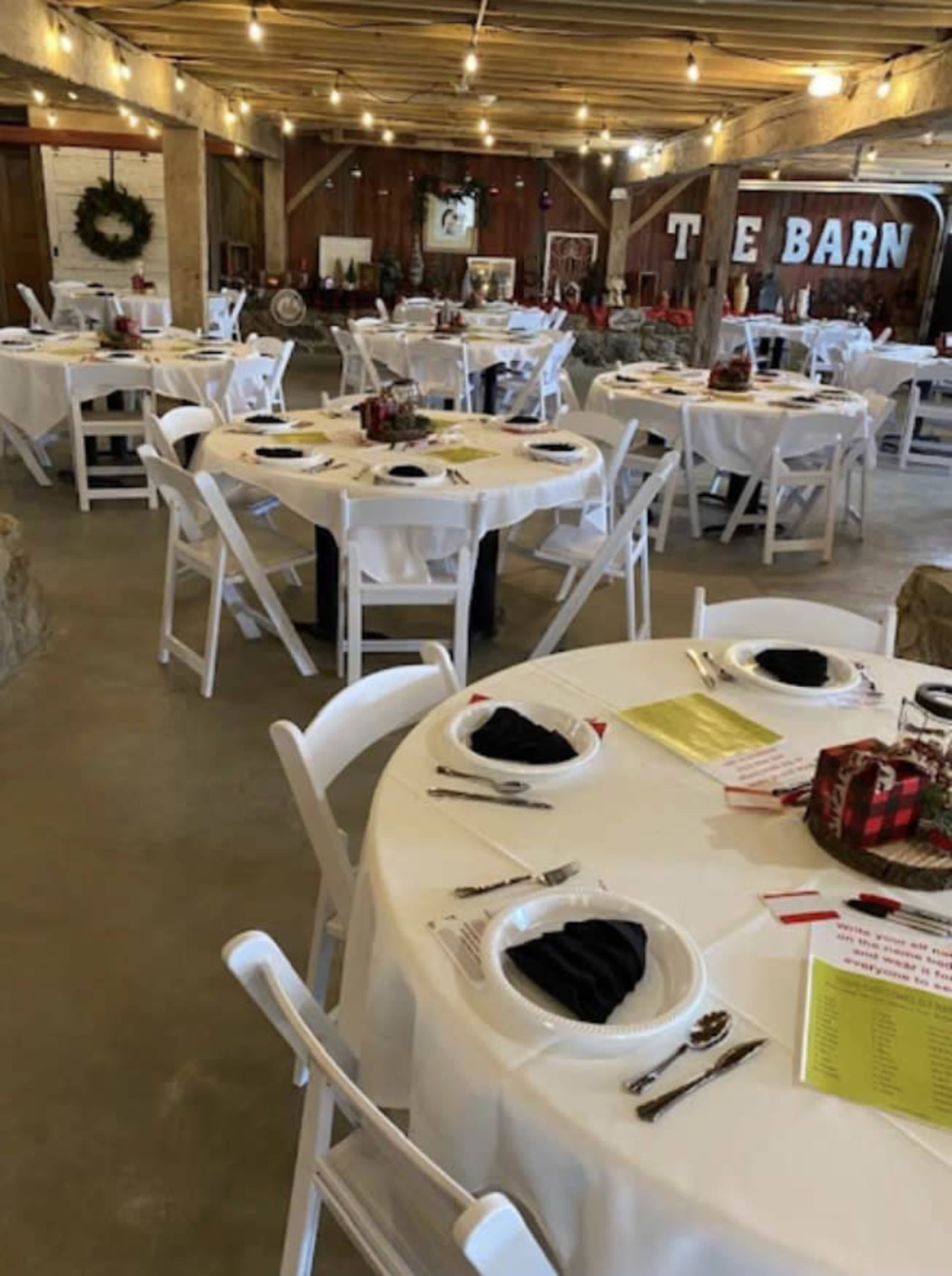 A decorated barn interior with several round tables set for a meal, each featuring white tablecloths and black napkins.