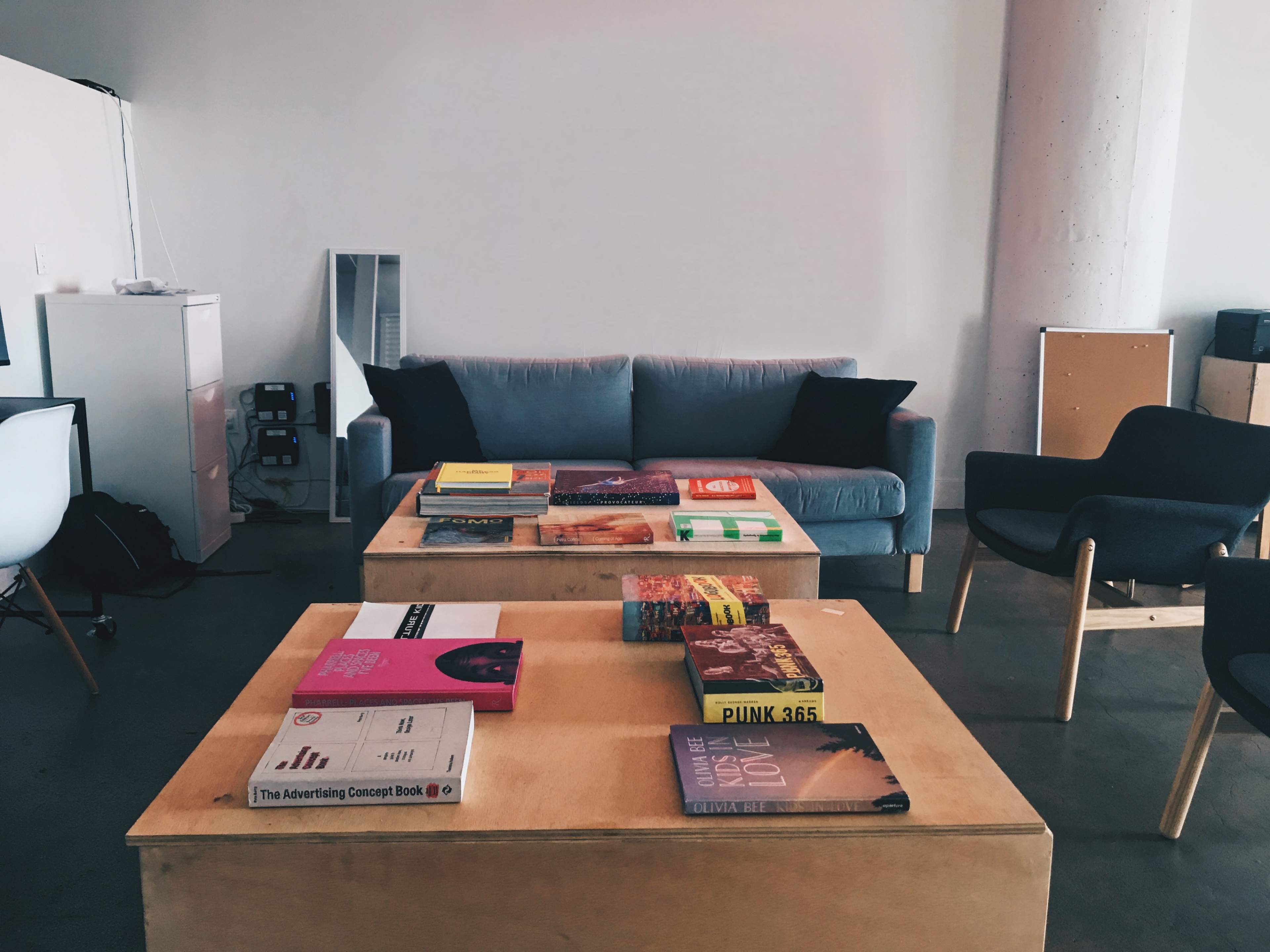 A living space features a gray couch and two black chairs arranged around wooden coffee tables covered with various books.