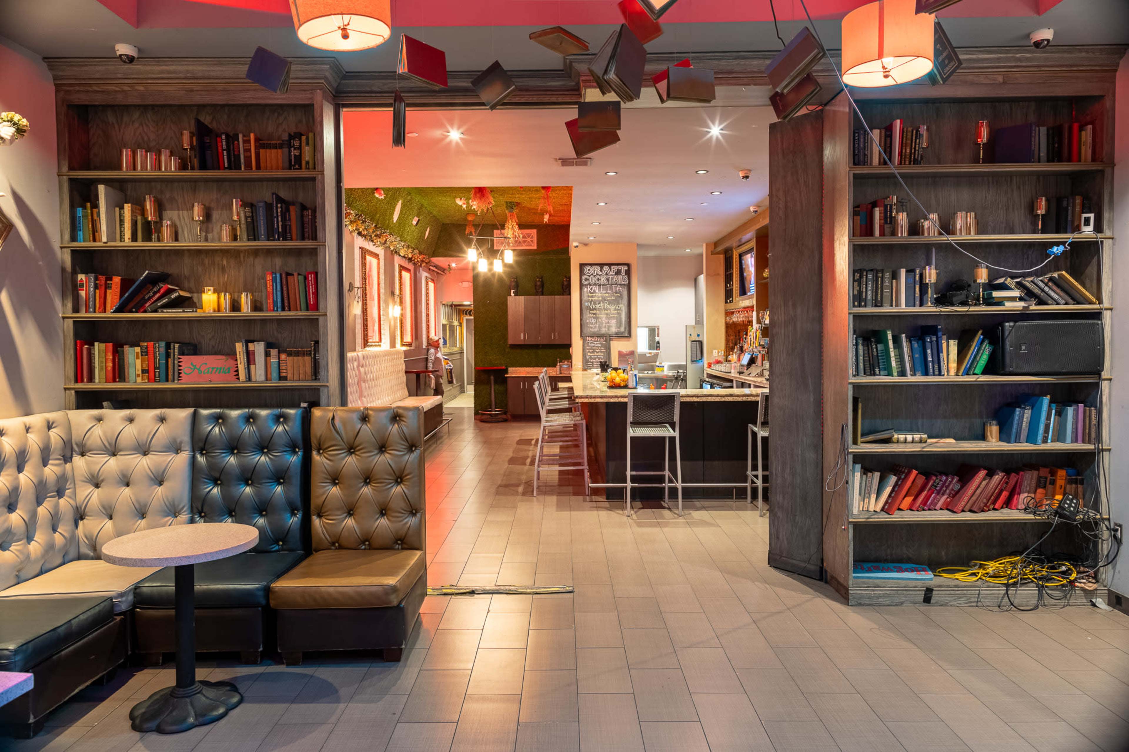 A cozy interior with a seating area featuring a tufted couch and a bookshelf filled with books, leading to an open space with a bar and a kitchen area.