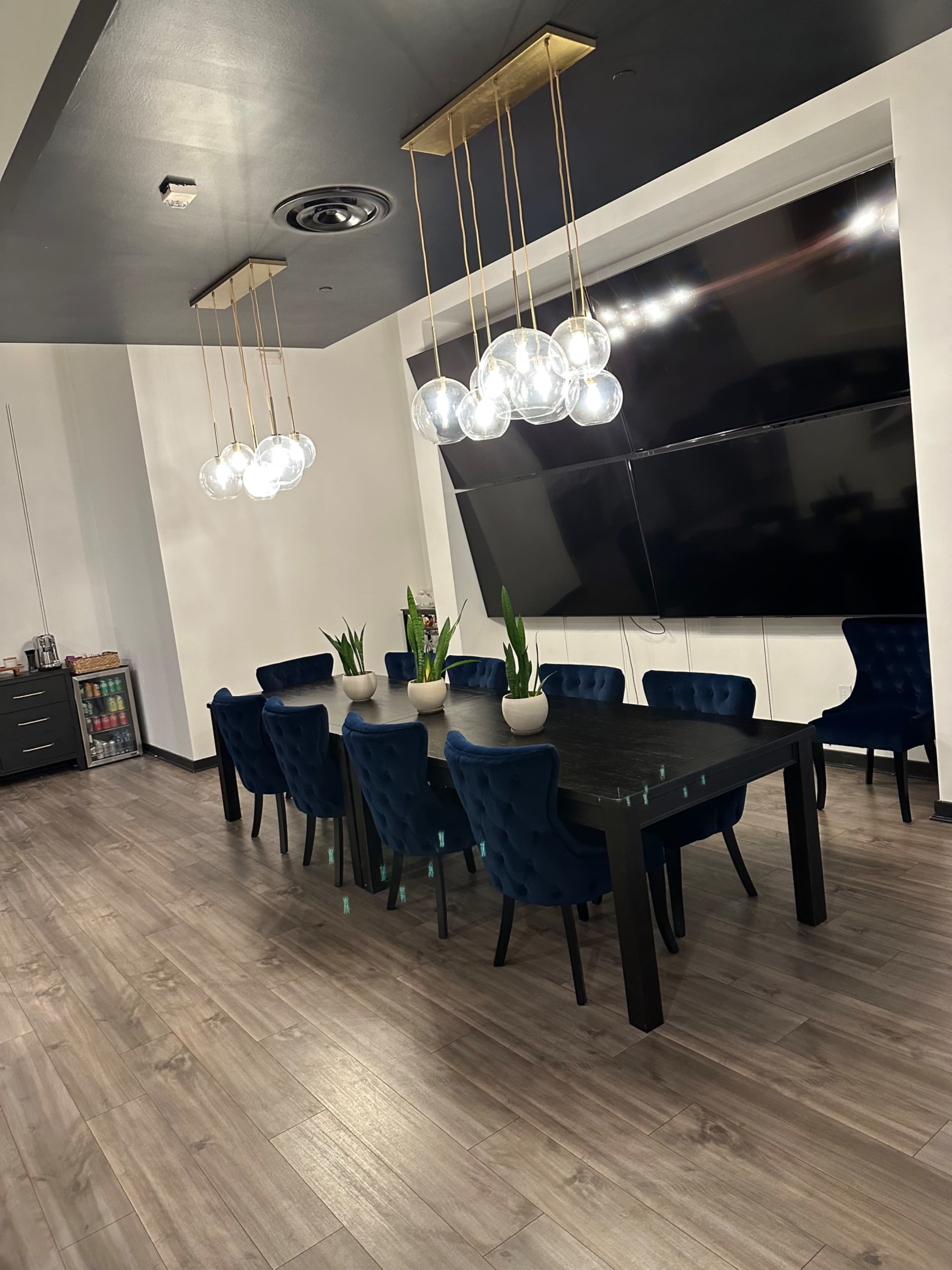 A modern dining area features a large black table surrounded by blue velvet chairs, with pendant lights hanging above and a large television mounted on the wall.