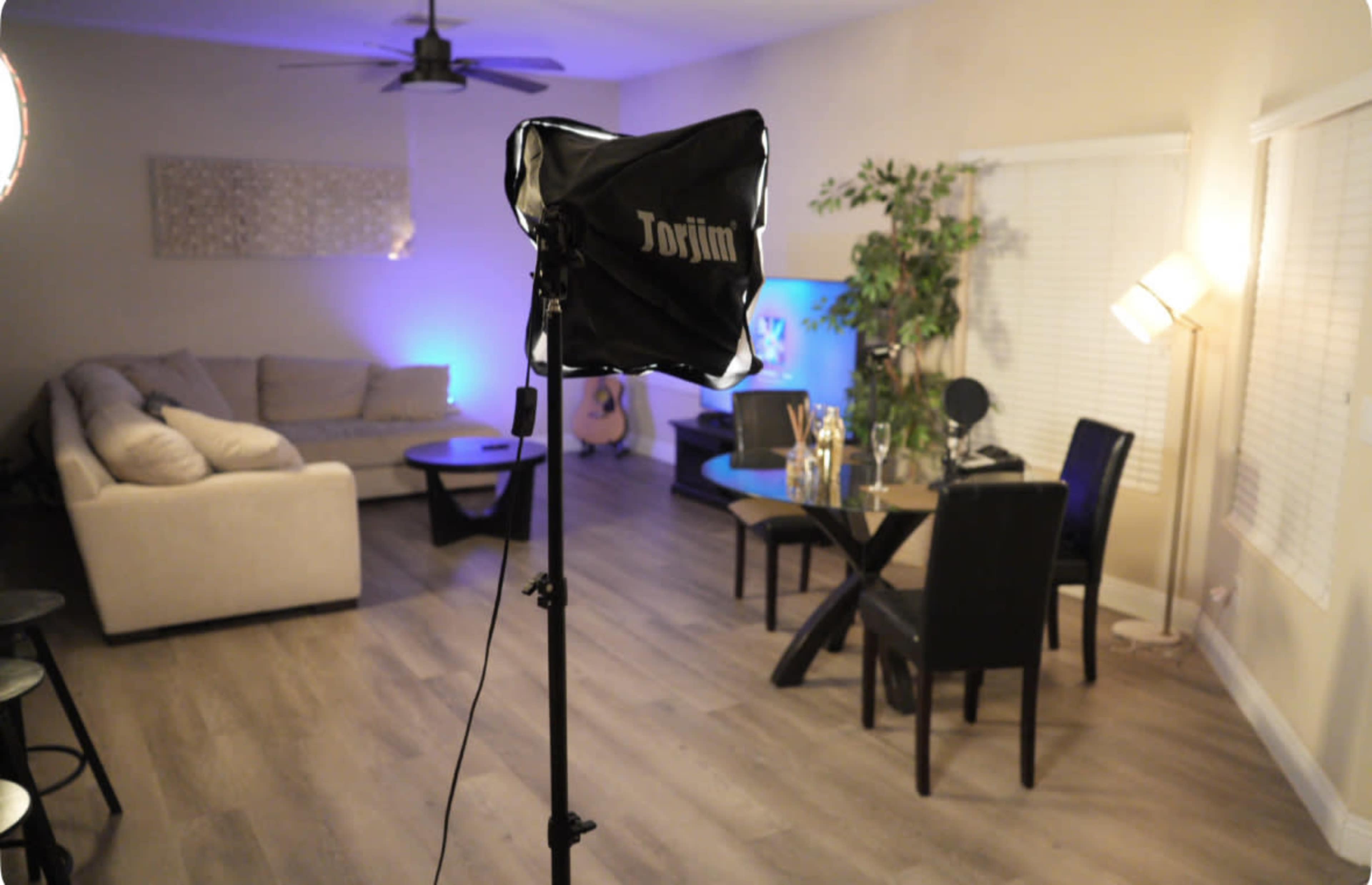 The image shows a well-lit living room with a couch, a coffee table, and a dining area featuring a plant and a guitar in the corner.