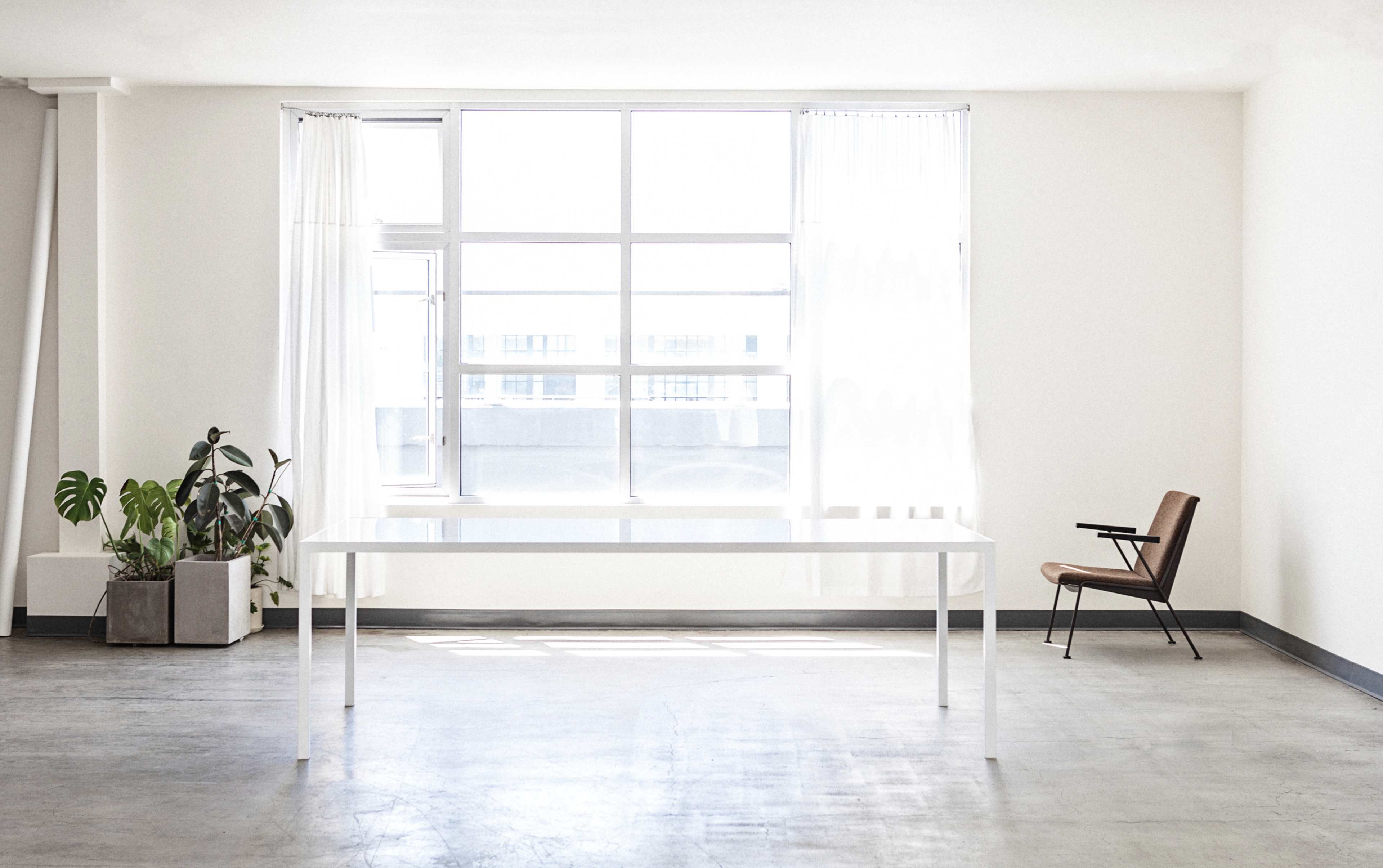 A bright, minimalistic room features a large window, a white table, a single chair, and a potted plant.
