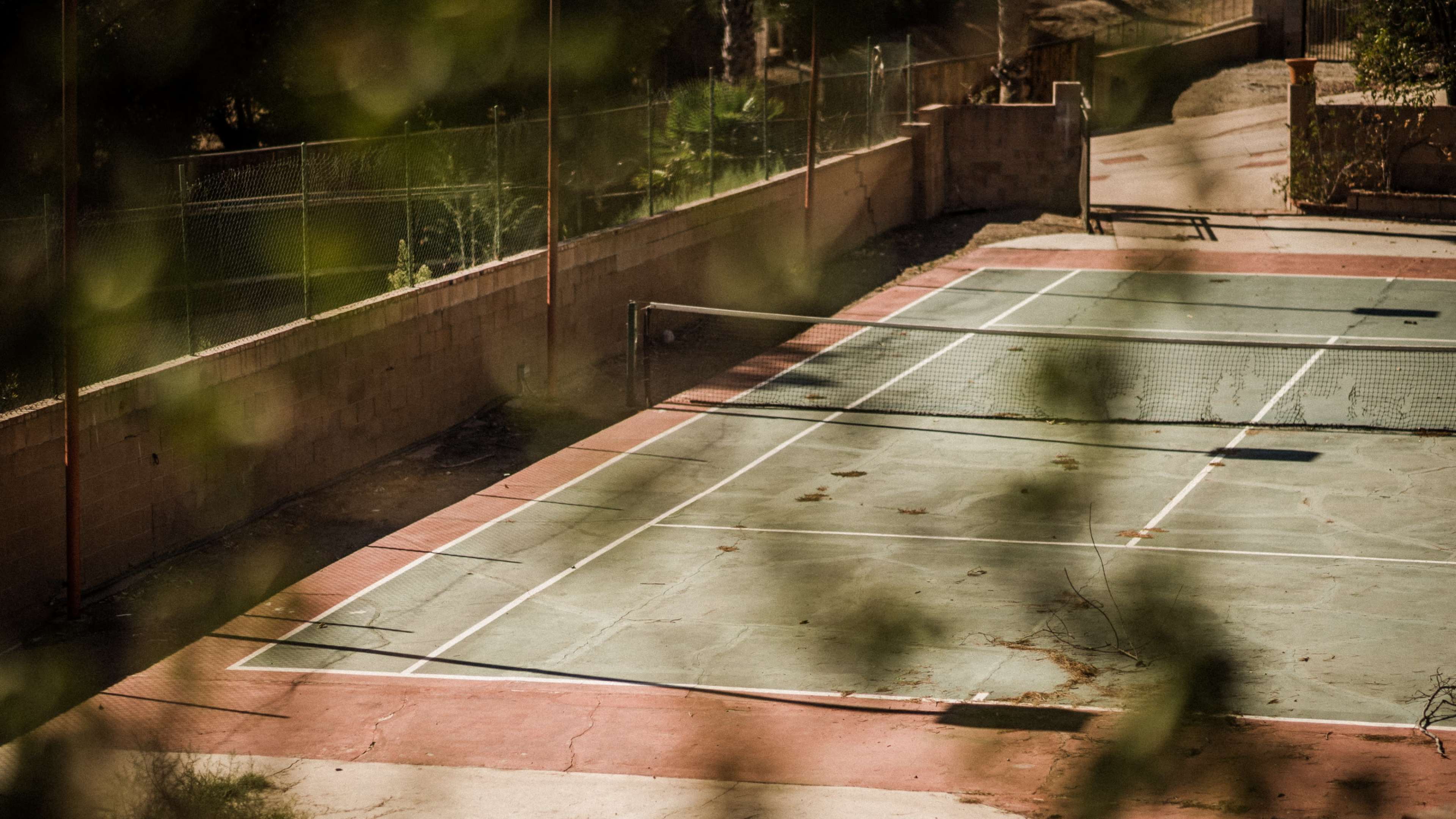 An empty tennis court with worn green and red surfaces is surrounded by a sparse landscape.